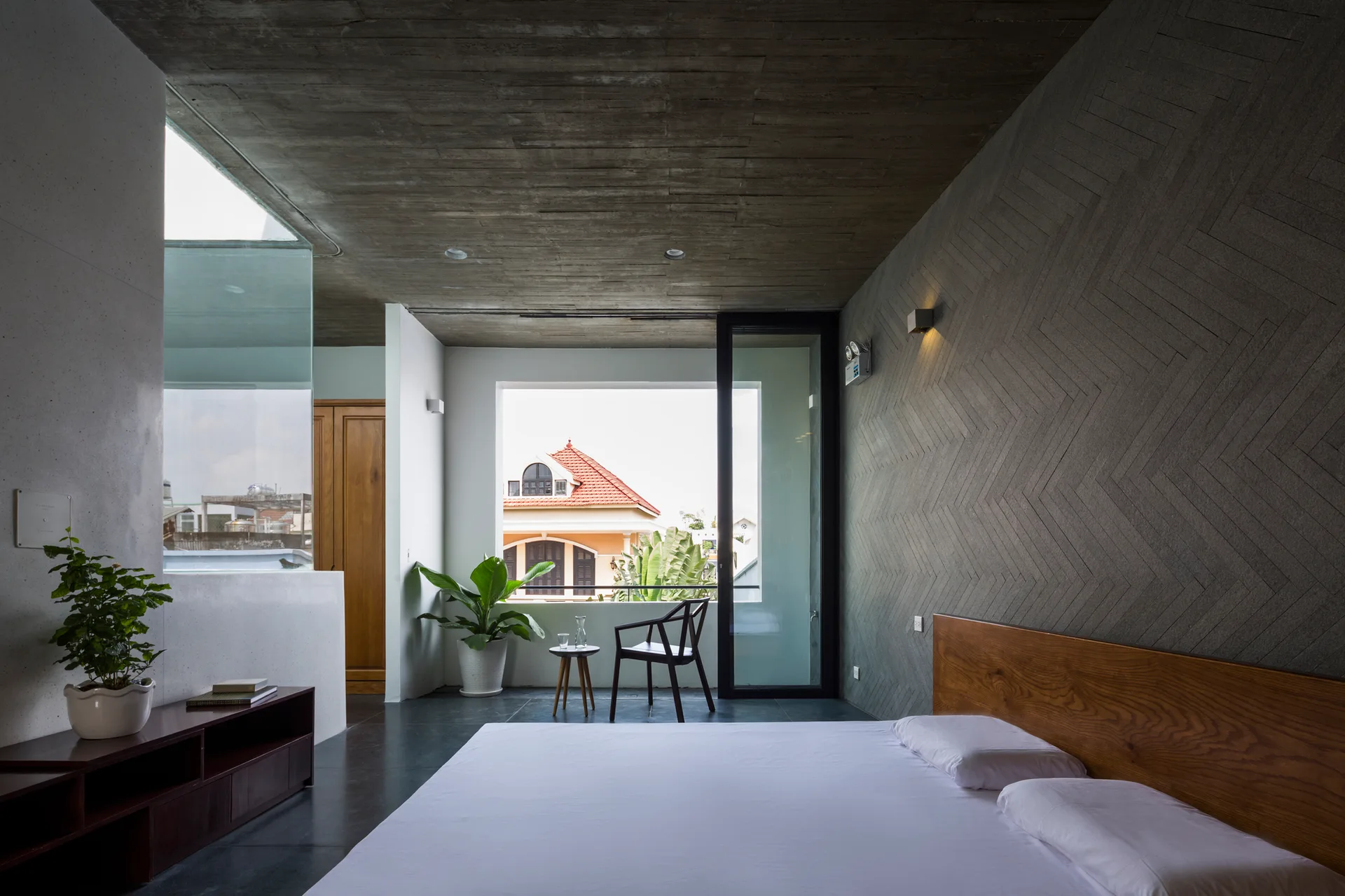 A modern bedroom features a textured grey herringbone wall, raw concrete ceiling, and low-profile wood bed near a large window.