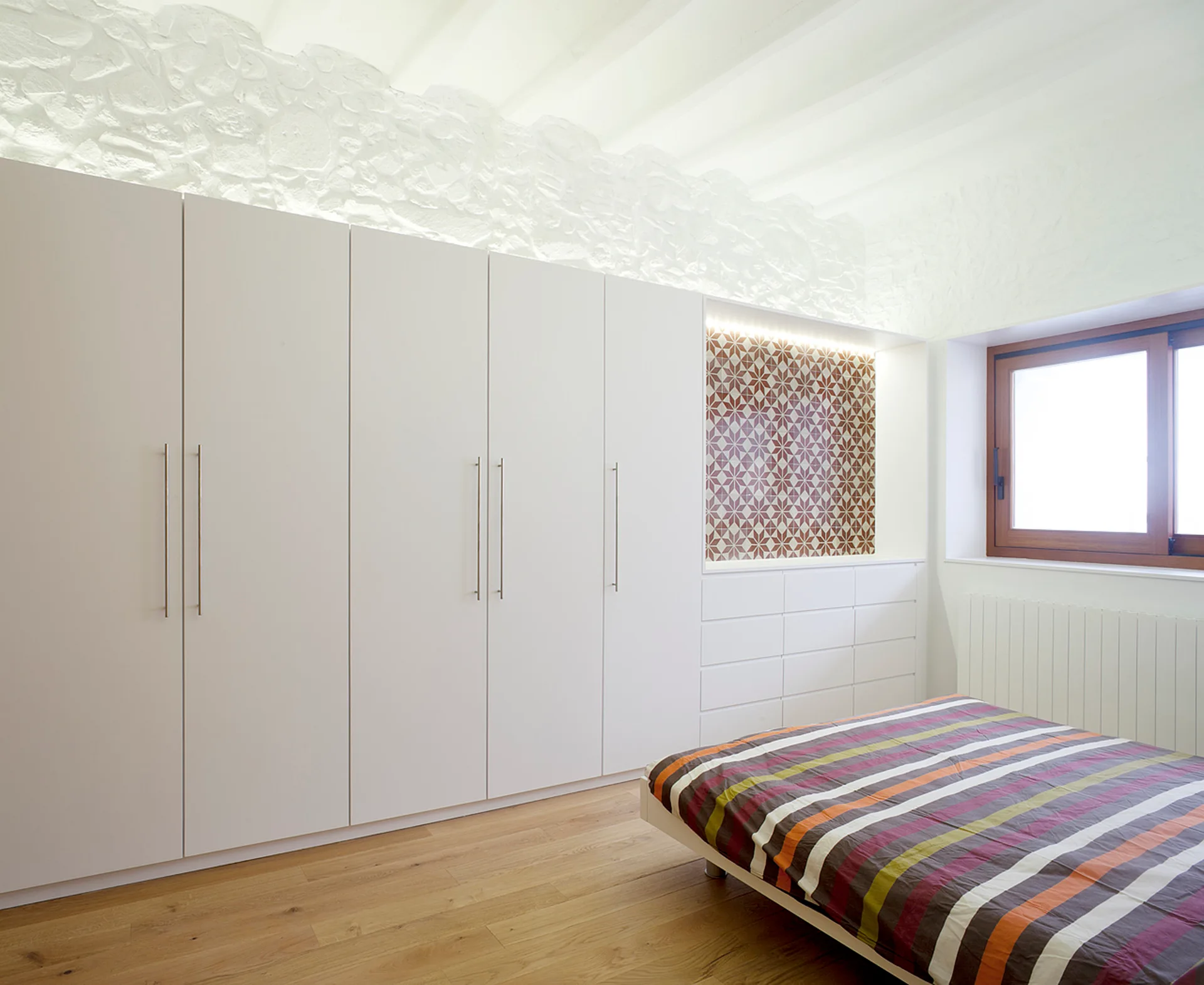A minimalist bedroom features a large white floor-to-ceiling wardrobe with a geometric tiled alcove next to a bed with striped bedding.