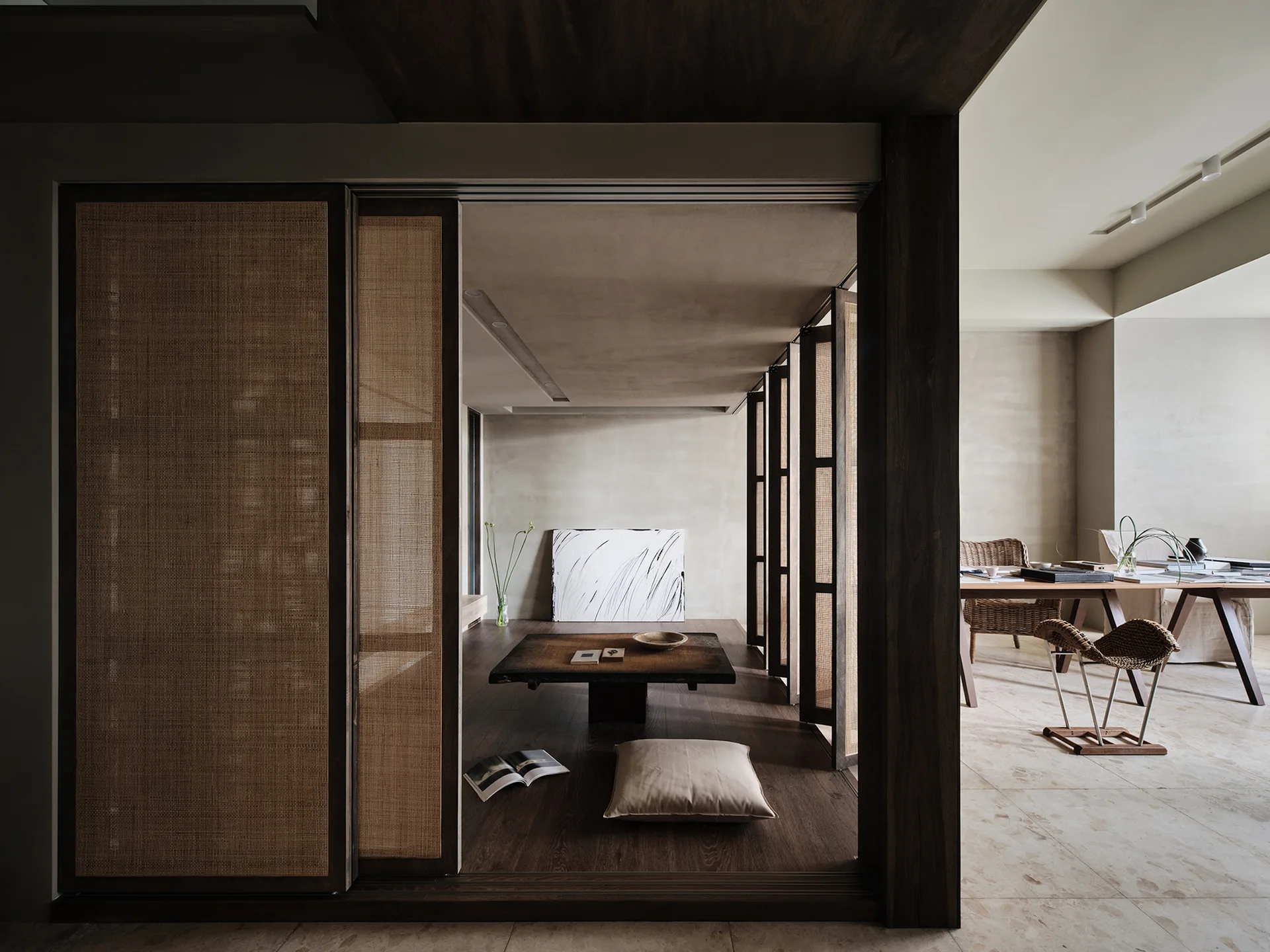 A minimalist Japandi interior featuring a raised wood platform with a low table separated from a dining area by woven rattan sliding screens.