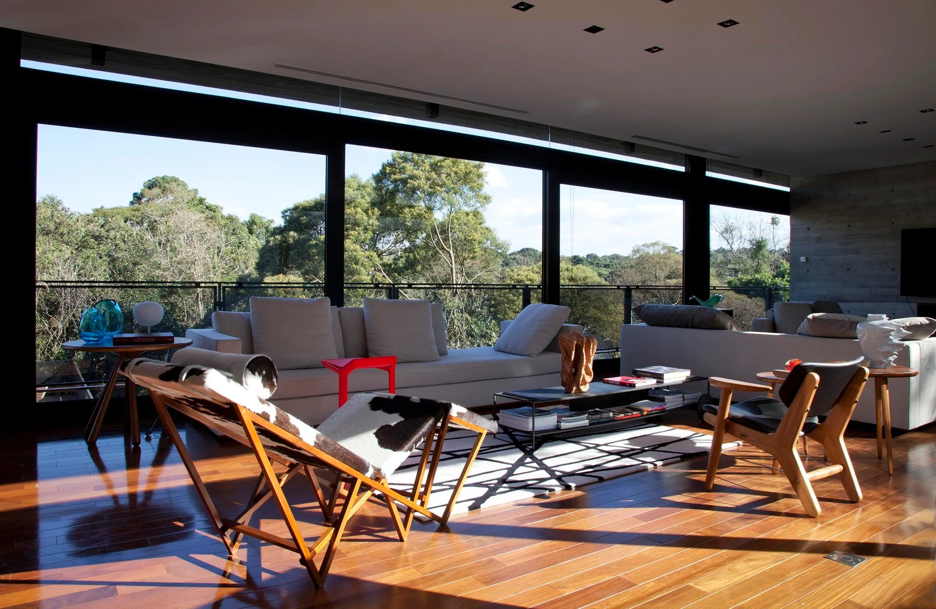 A modern living room features a cream sofa, a graphic line rug, wood-framed cowhide chairs, and a wall of floor-to-ceiling windows.