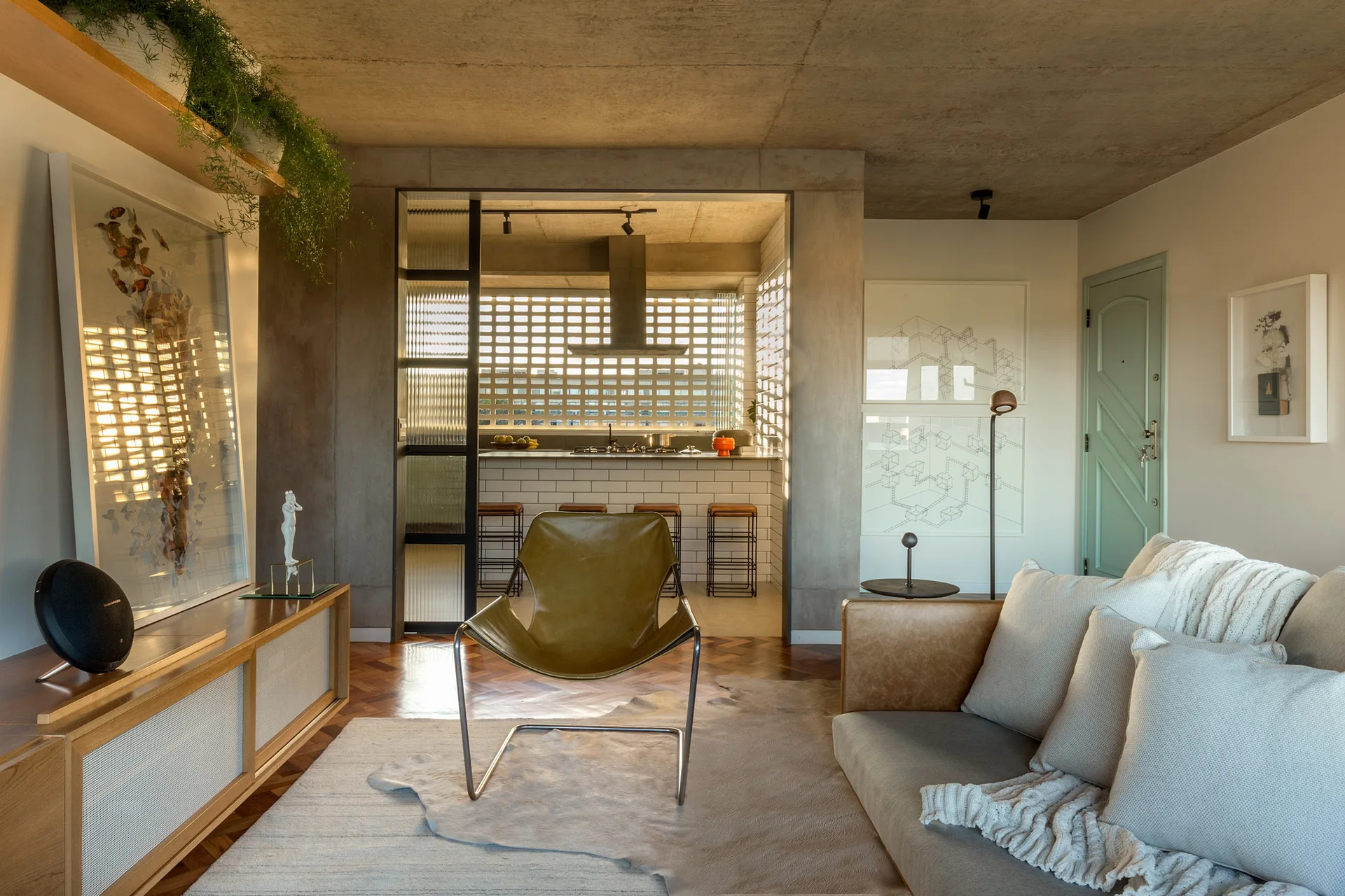 A modern living room features an olive leather chair, herringbone wood floors, and an open view into a kitchen with a white breeze block wall.