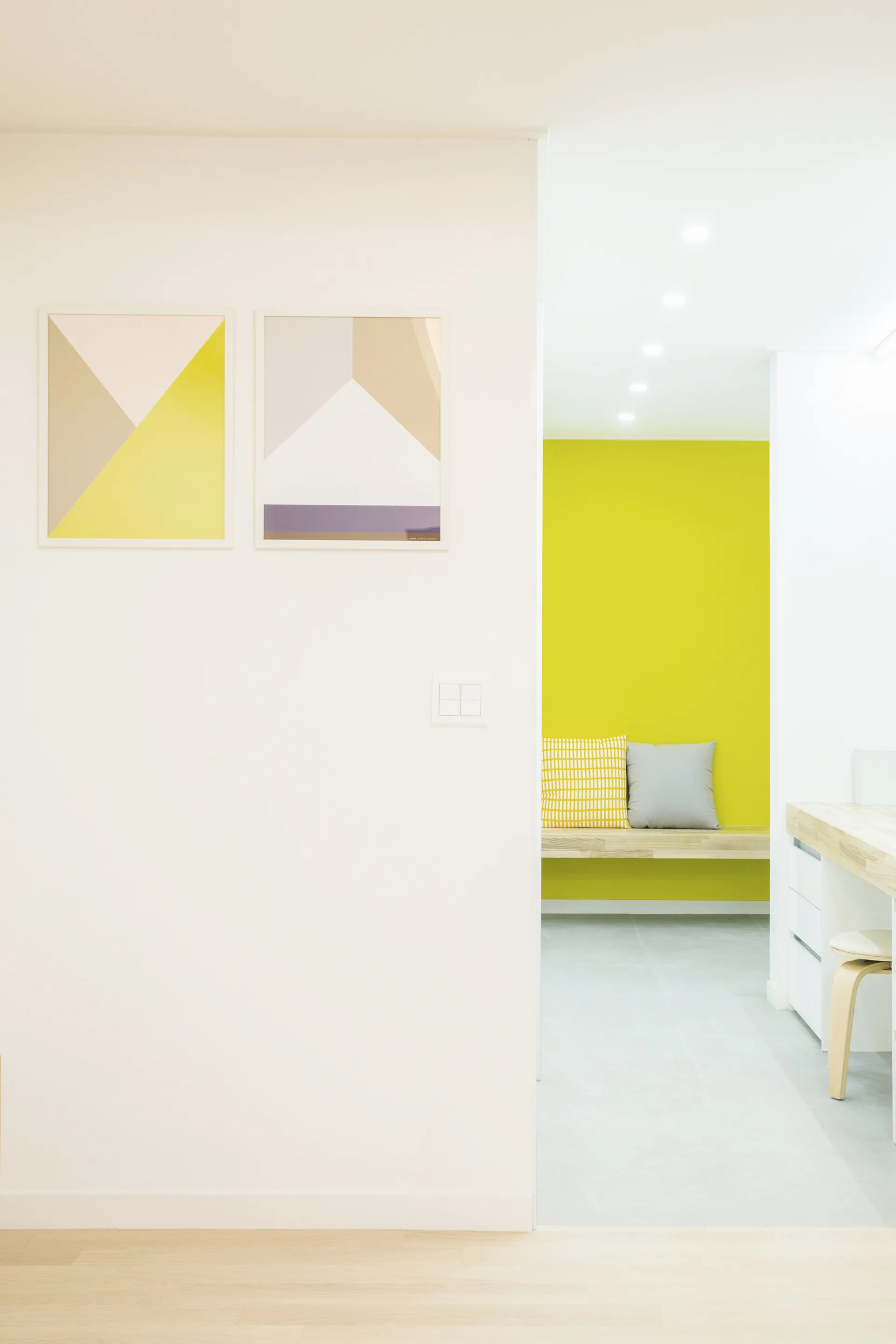 Two framed geometric prints hang on a white wall in a hallway leading to a lime green accent wall with a wooden bench.
