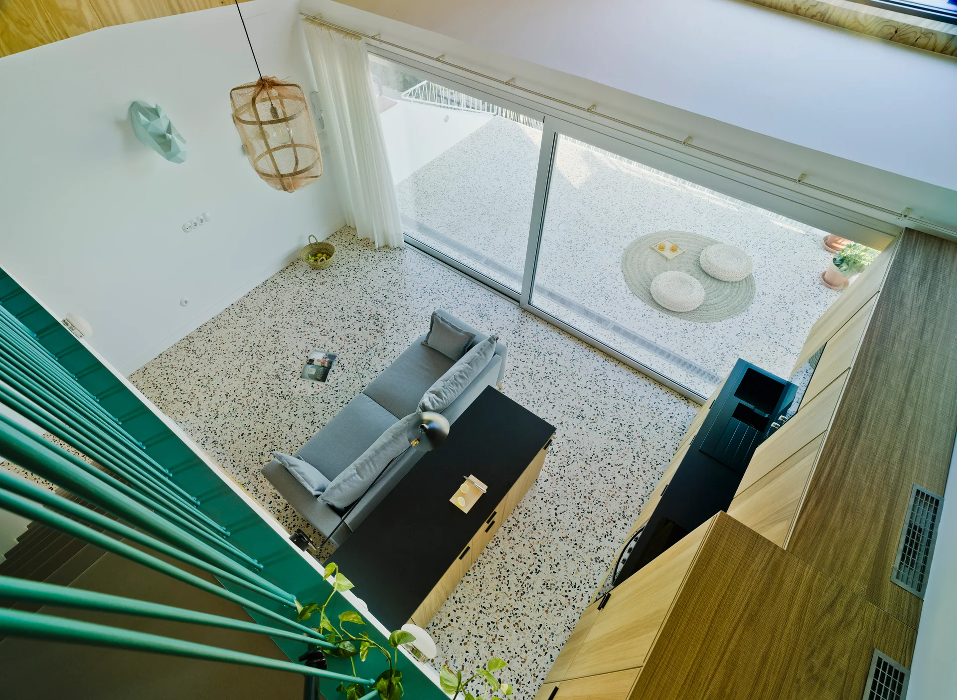 High-angle view of a modern living room featuring a grey sofa on speckled terrazzo flooring next to wood kitchen cabinets and a teal staircase railing.