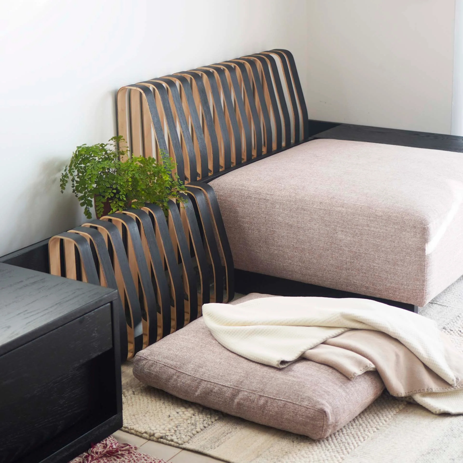 A modern low-profile upholstered sofa features a bent-wood slatted backrest alongside a matching floor cushion and a black wooden side table.