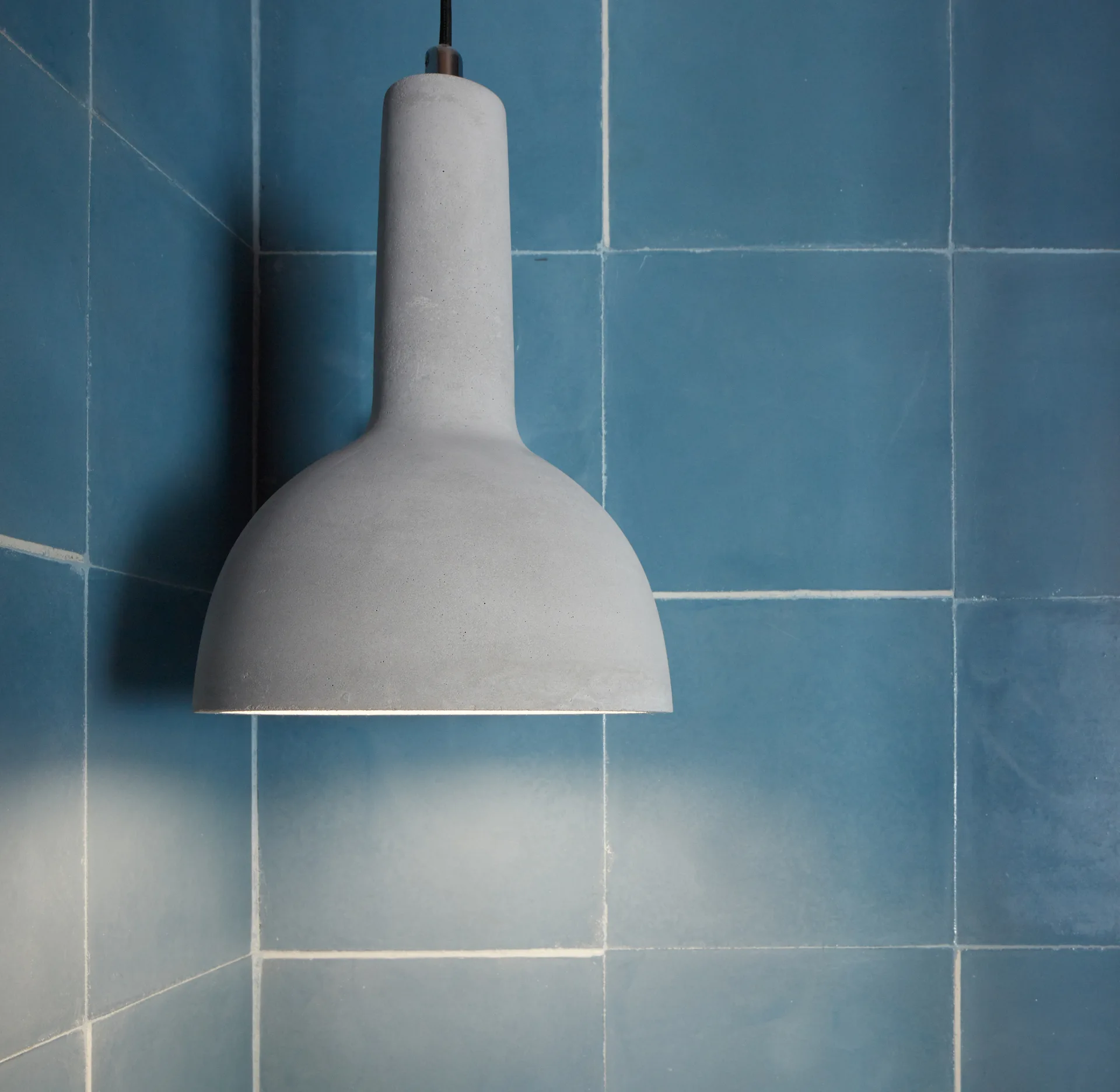 A matte grey concrete pendant light hangs in a corner against a wall of square blue tiles with white grout lines.