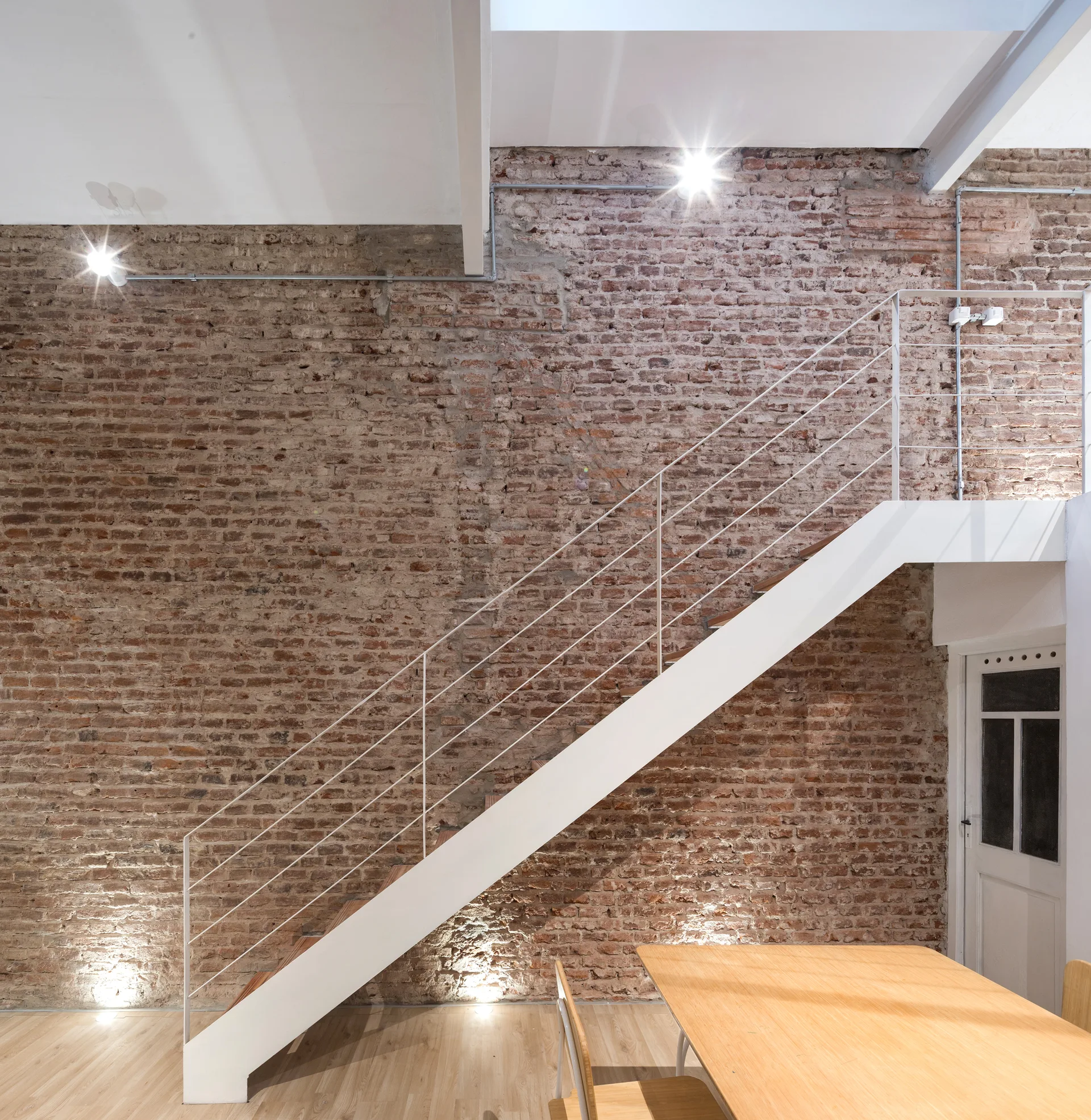 A modern white metal staircase ascends along a large exposed brick wall next to a light wood dining table and wood flooring.