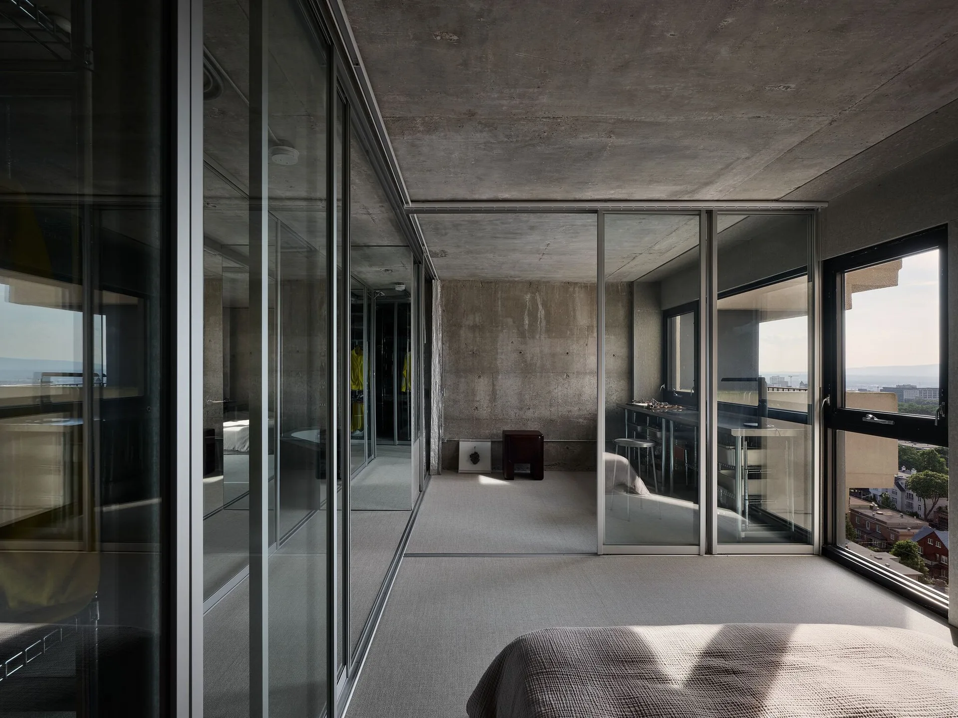 A minimalist bedroom featuring raw exposed concrete walls and ceiling, sliding glass partitions, and a grey carpeted floor.
