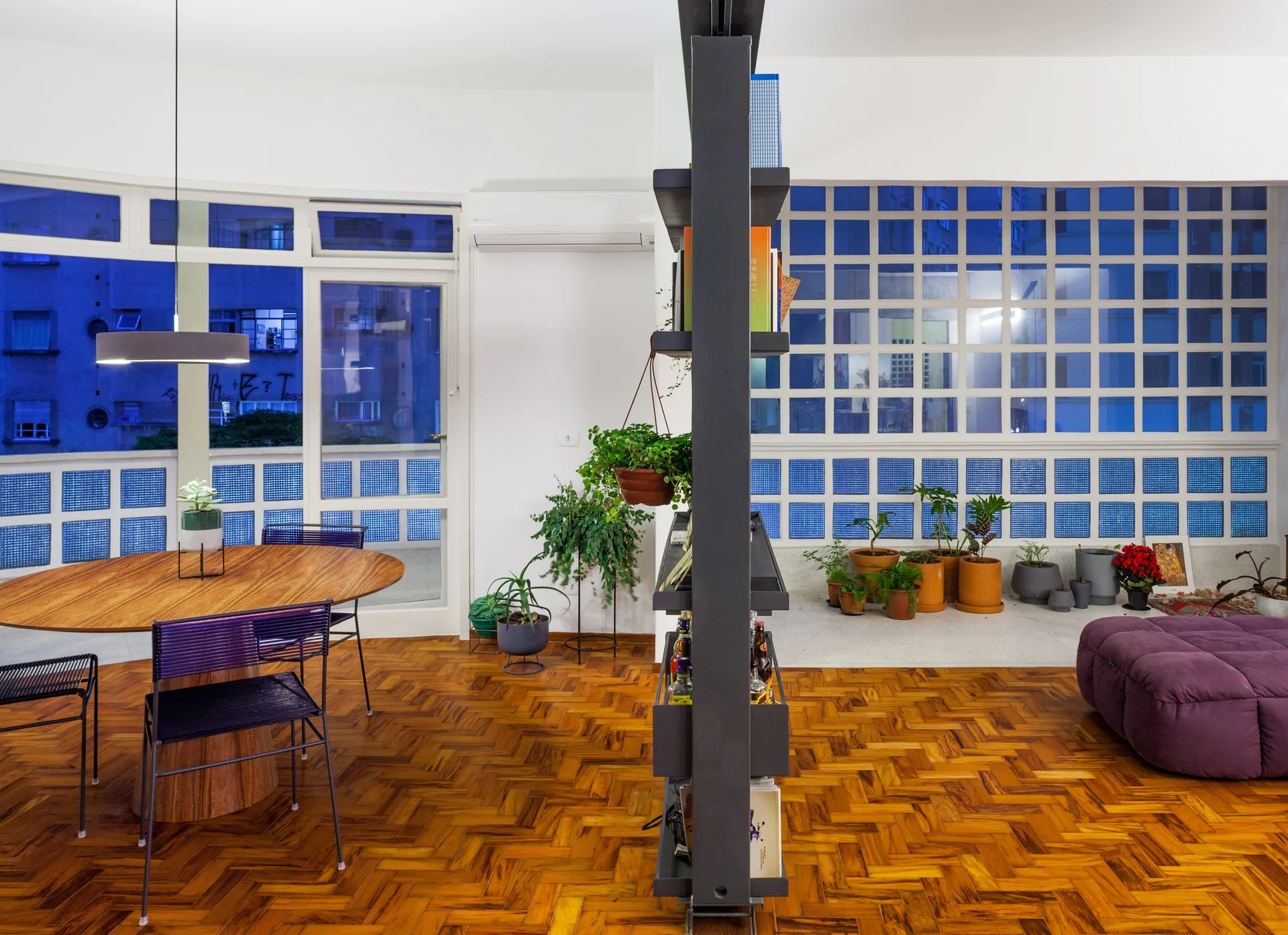 An open plan apartment interior featuring a herringbone wood floor, a central dark shelving unit, a dining table, and blue glass block windows.