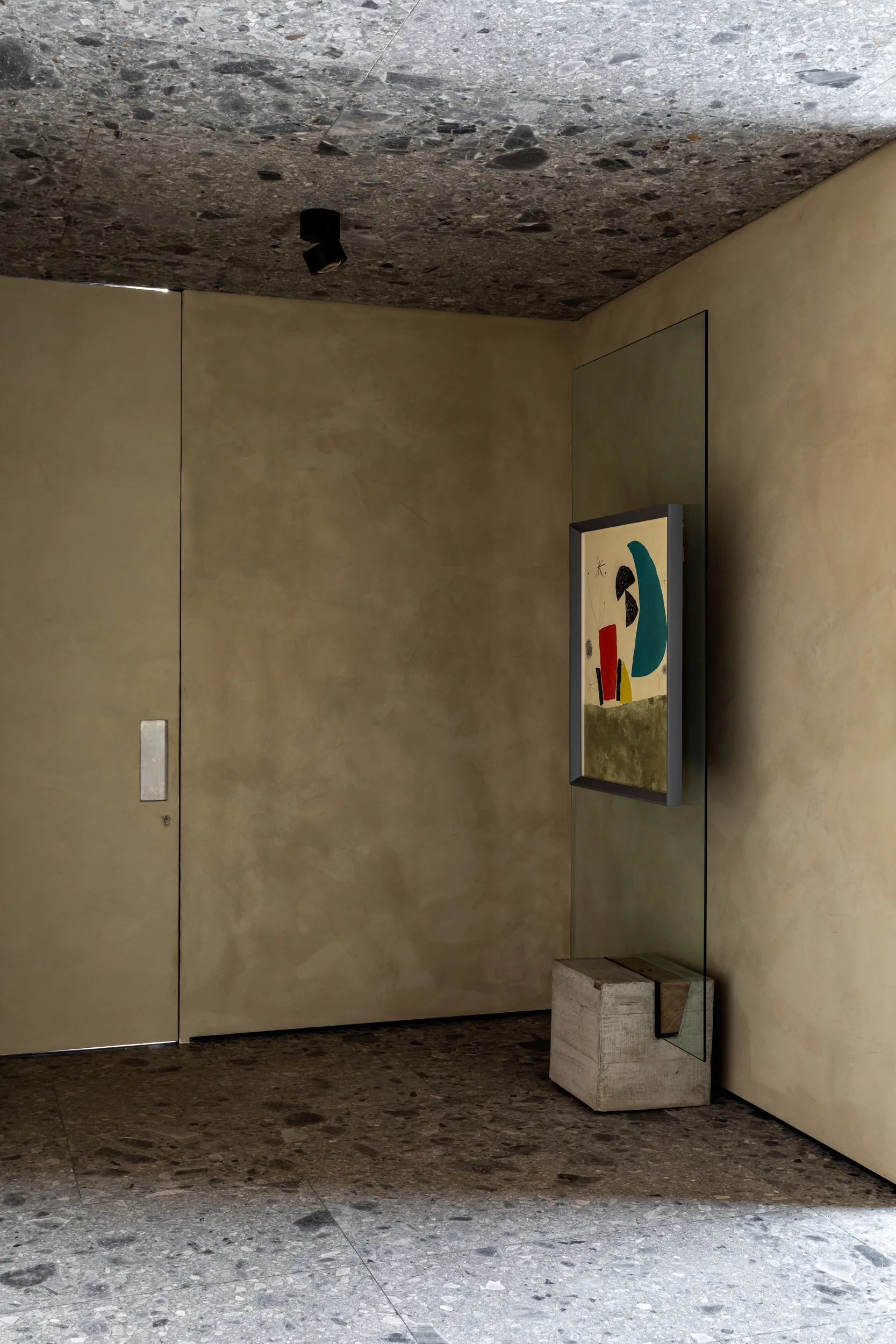 A minimalist entryway corner features a terrazzo floor and ceiling, matte tan plaster walls, and an abstract painting mounted on a glass partition.