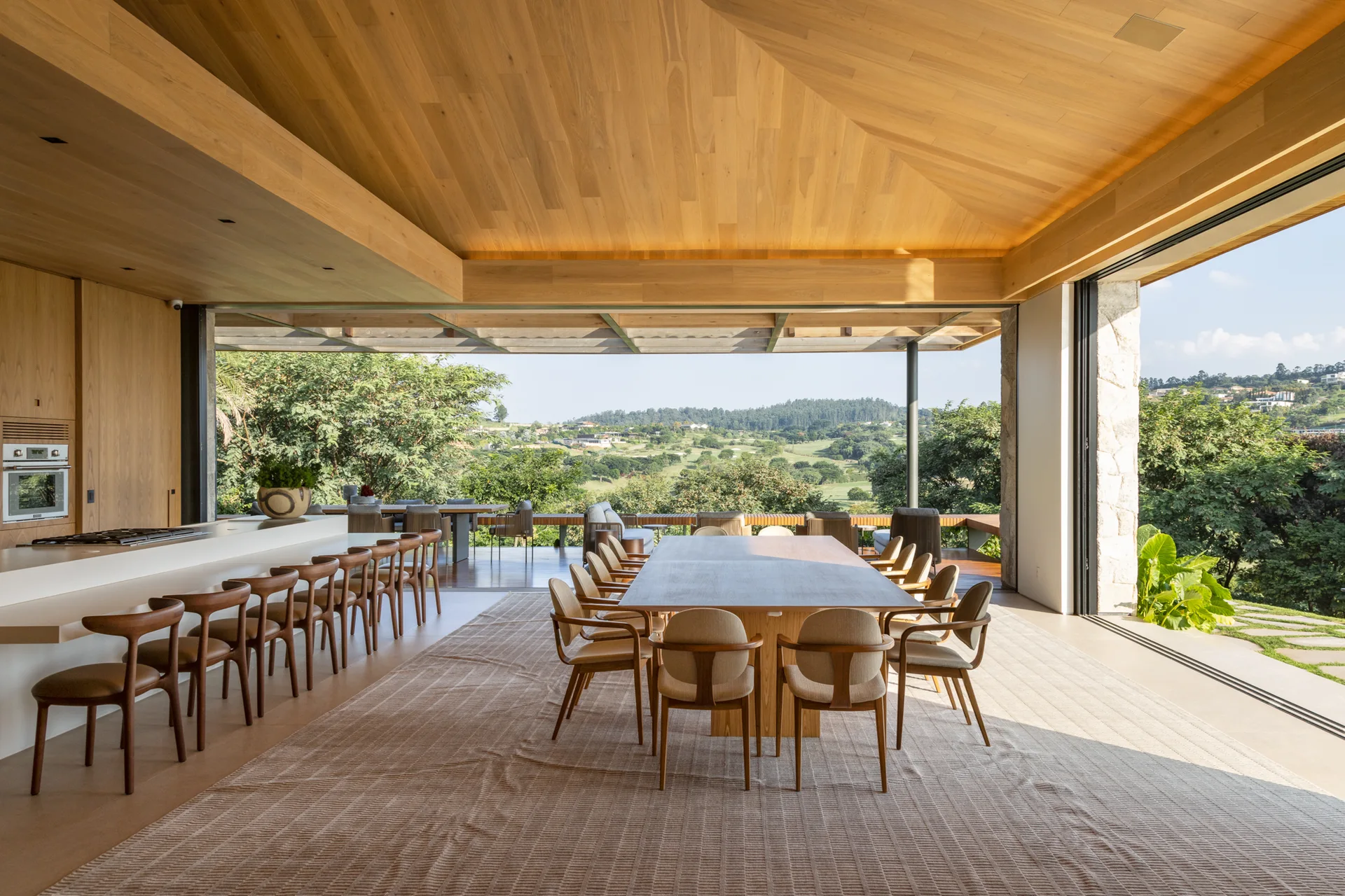 A large wooden dining table and chairs sit under a vaulted wood plank ceiling in an open-concept room featuring sliding glass walls with outdoor views.