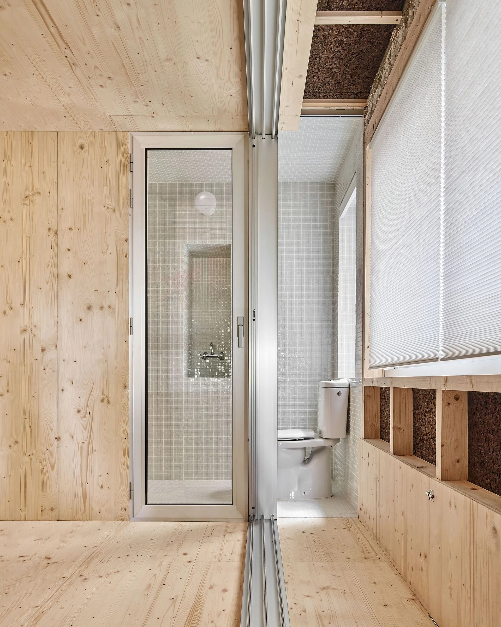 A modern bathroom features light-toned plywood walls next to a shower and toilet area tiled in white mosaic.