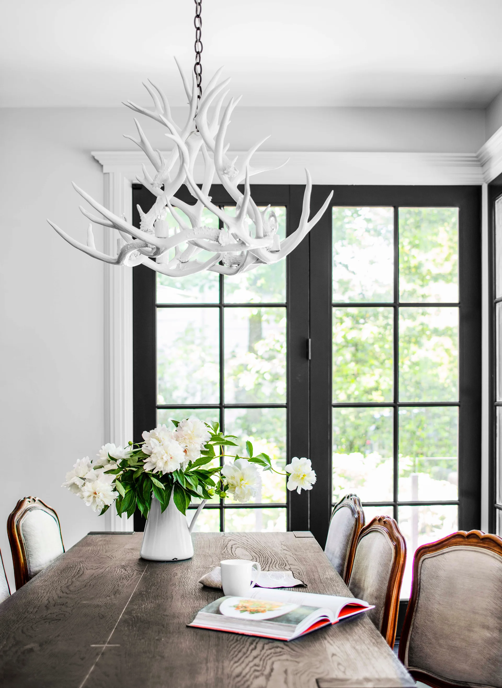 A dining room features a dark wood table with white peonies under a white antler chandelier and in front of black-framed doors.