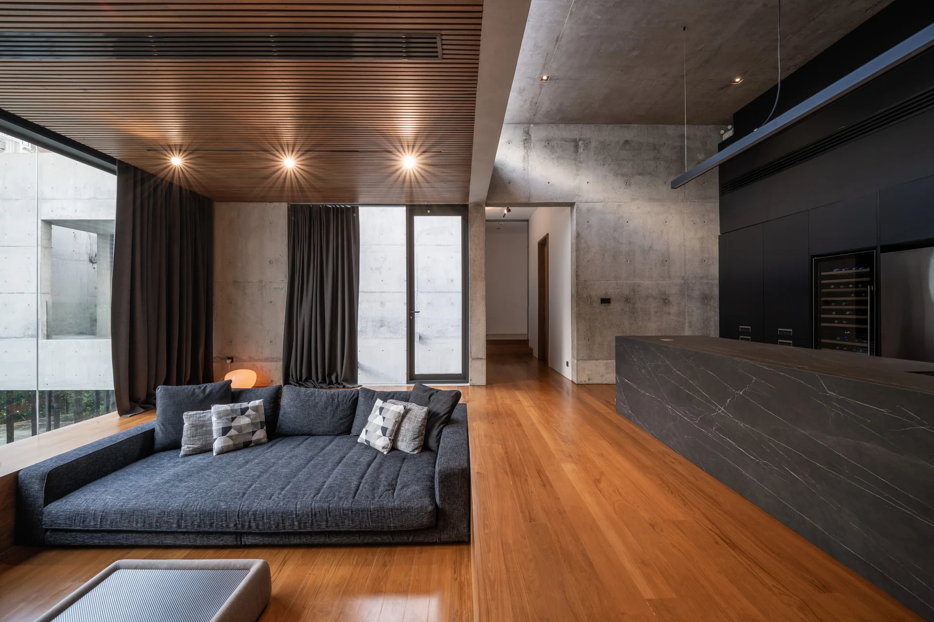 An open-plan living room featuring a low-profile grey sofa, warm wood flooring, raw concrete walls, and a linear wood slatted ceiling.
