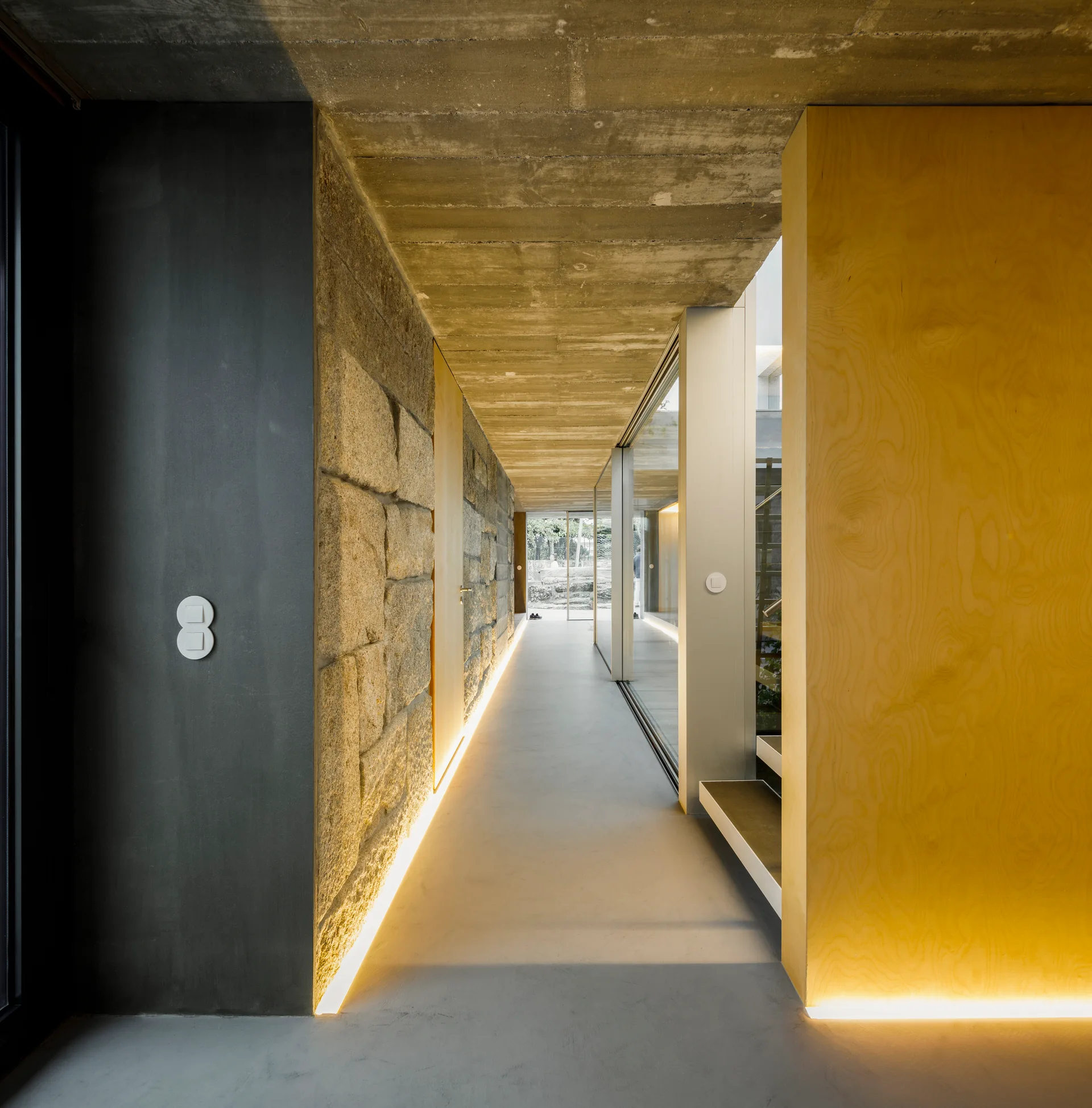 A minimalist hallway features an exposed concrete ceiling, a rugged stone wall, and integrated LED floor lighting casting a warm glow.