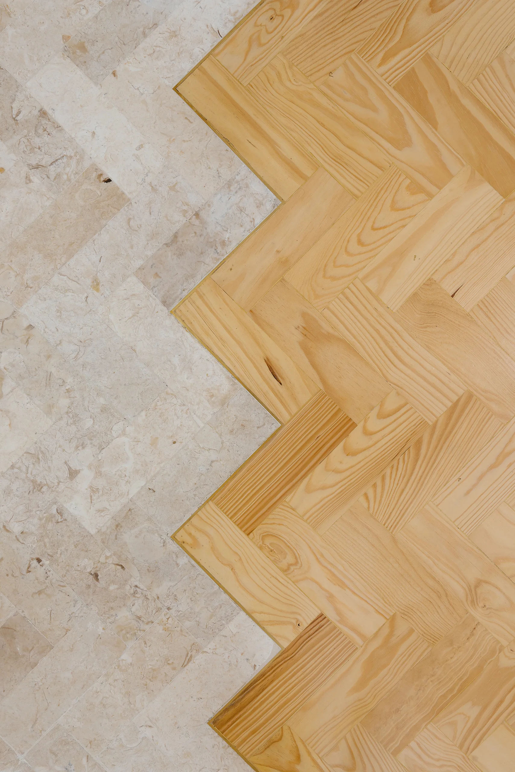 A close-up overhead view of a floor transition where beige stone tiles meet light natural wood planks in a shared herringbone pattern.