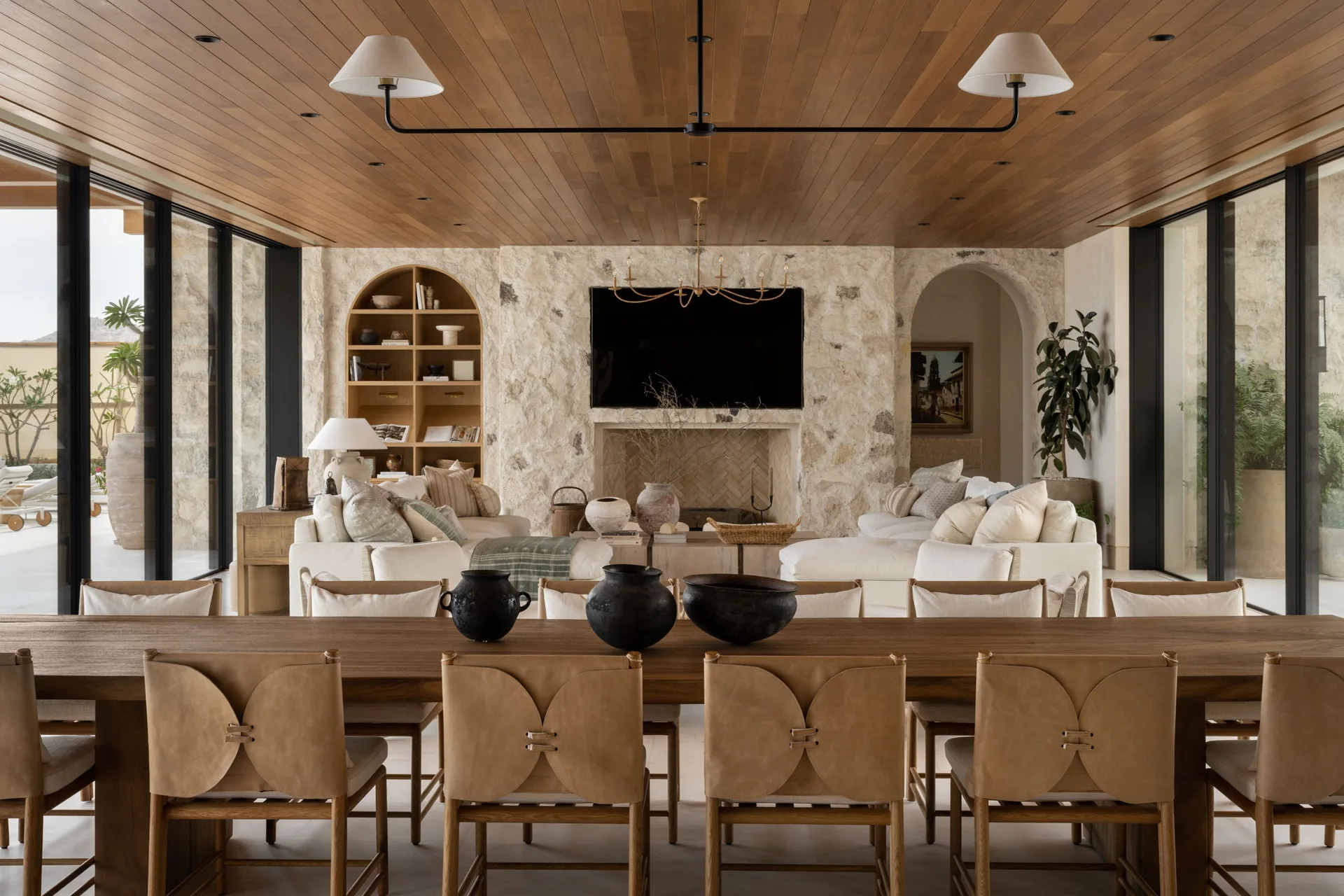 An open-plan dining and living room featuring a wood plank ceiling, a textured stone fireplace wall, and a long wooden table with leather-backed chairs.