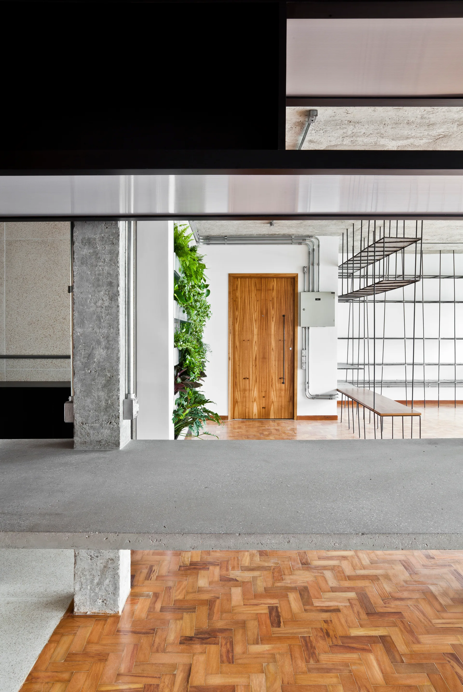 An industrial interior features a grey concrete countertop, a raw concrete column, warm herringbone parquet wood floors, and a vertical garden wall.