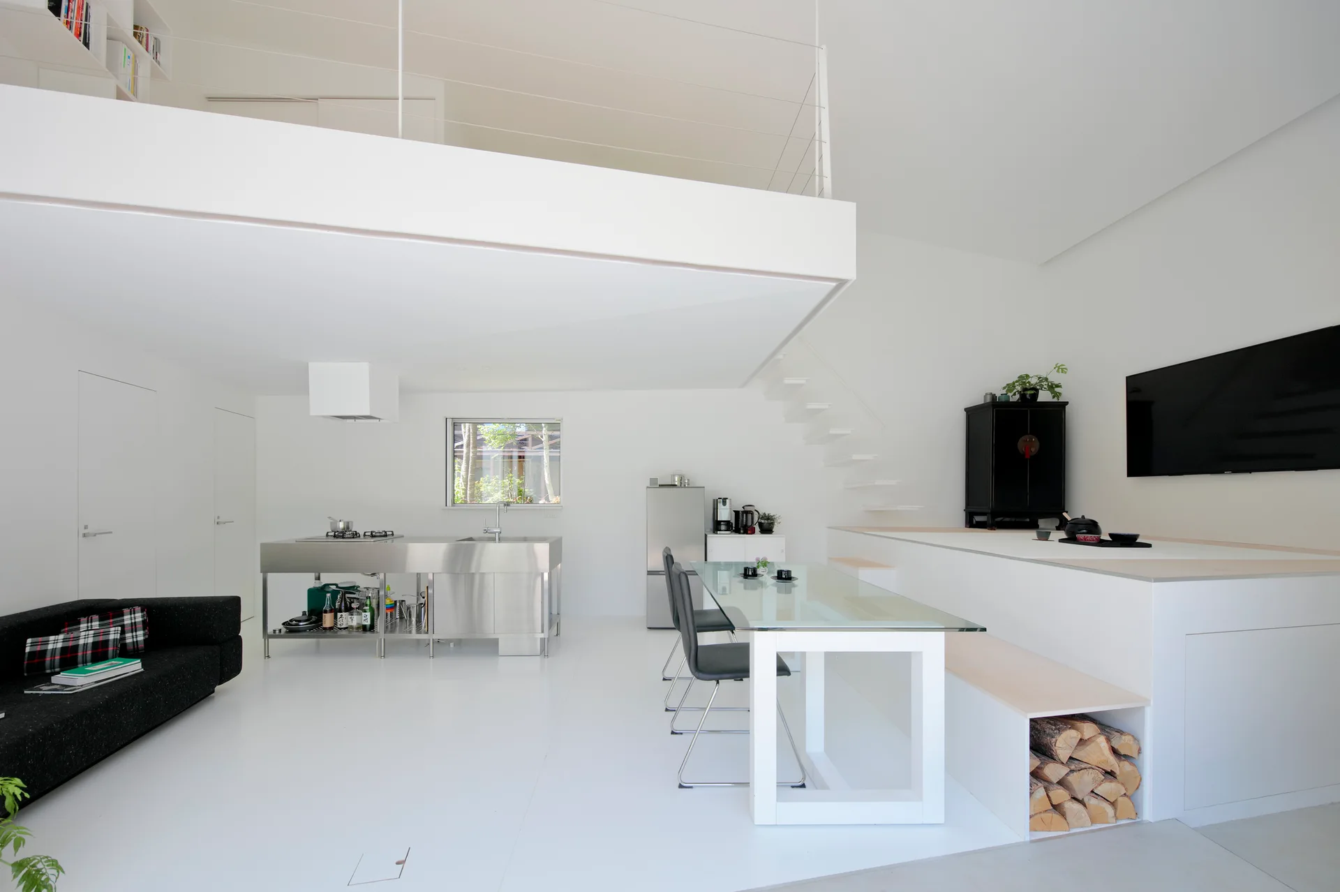 An open-concept white interior features a stainless steel kitchen island, a glass dining table, and a staircase leading to a mezzanine loft.
