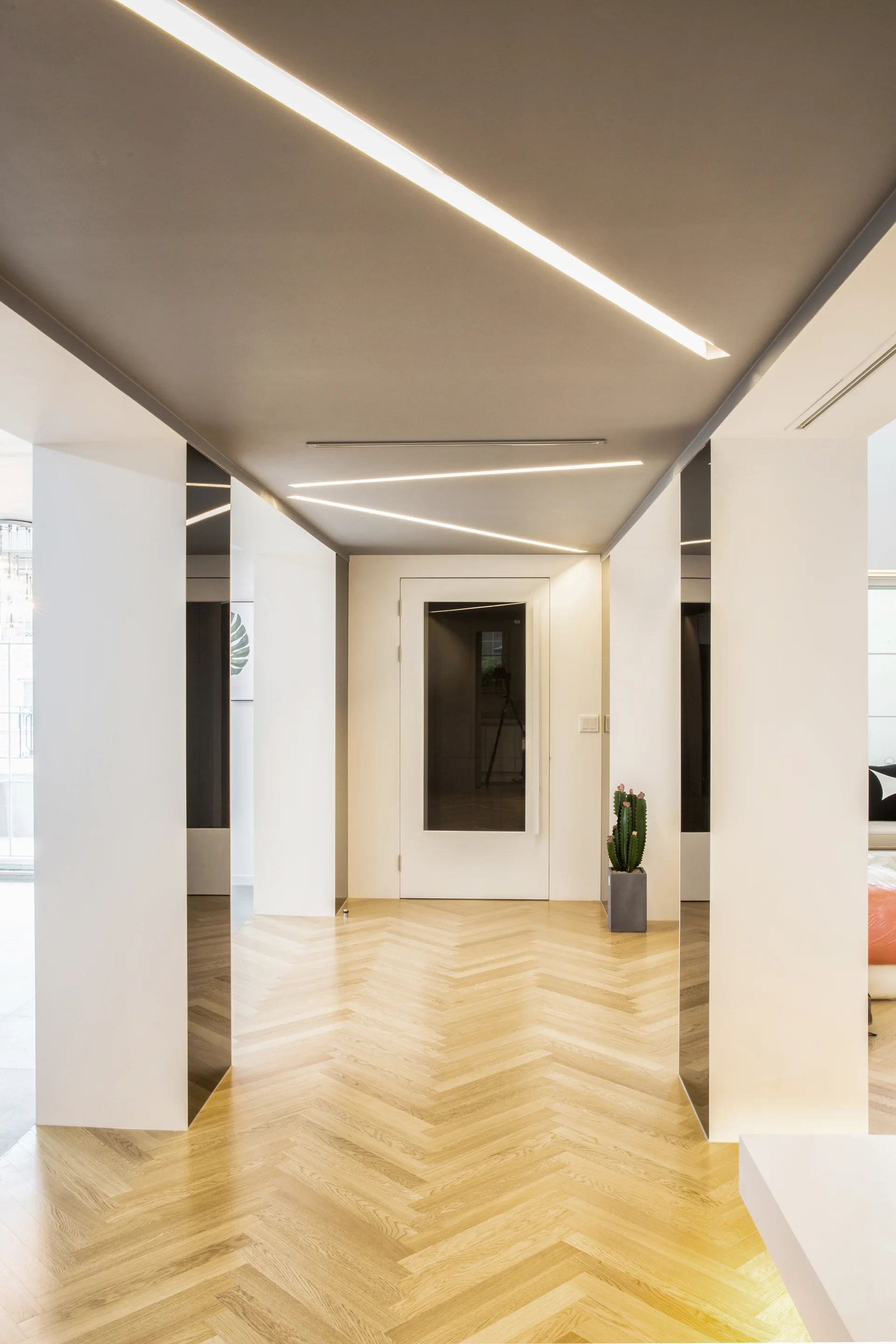 A modern hallway features light wood herringbone flooring, white walls, and a grey ceiling with bright recessed linear light strips.
