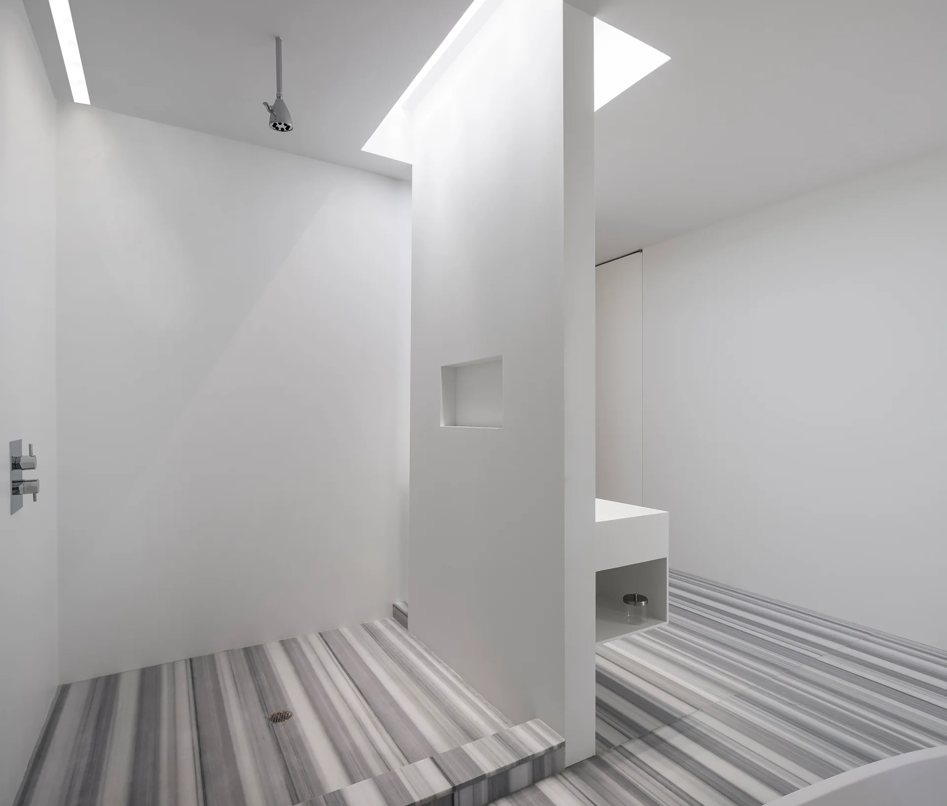 A minimalist white bathroom features a central partition wall, a ceiling shower head, and grey striped marble floor tiles.