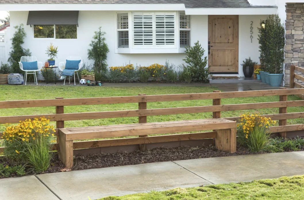 A slatted wood bench sits against a horizontal split-rail fence in a landscaped yard in front of a white house with blue patio chairs.