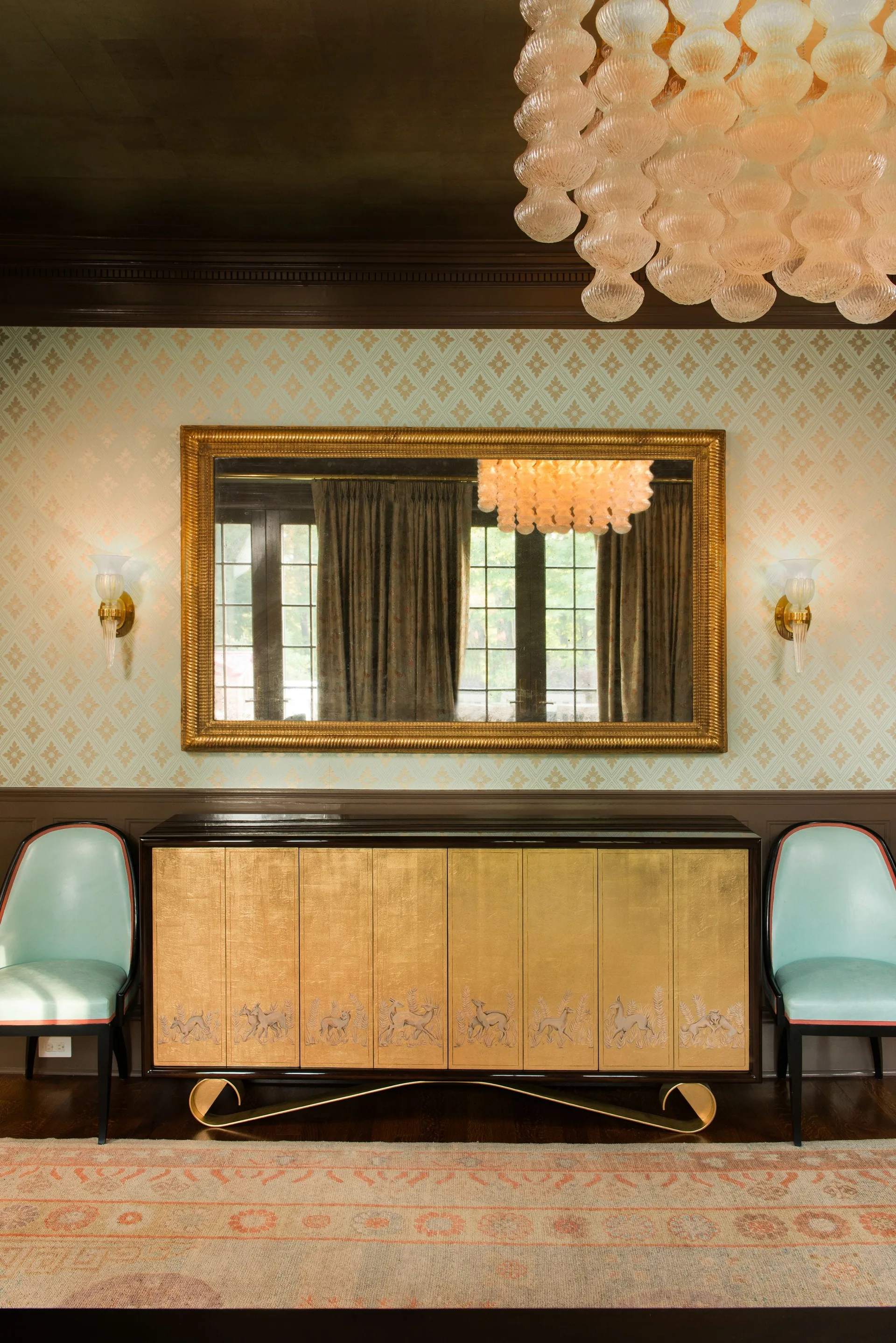 A gold-fronted sideboard flanked by two light blue chairs sits beneath a large gilded mirror on a geometric patterned wallpapered wall.