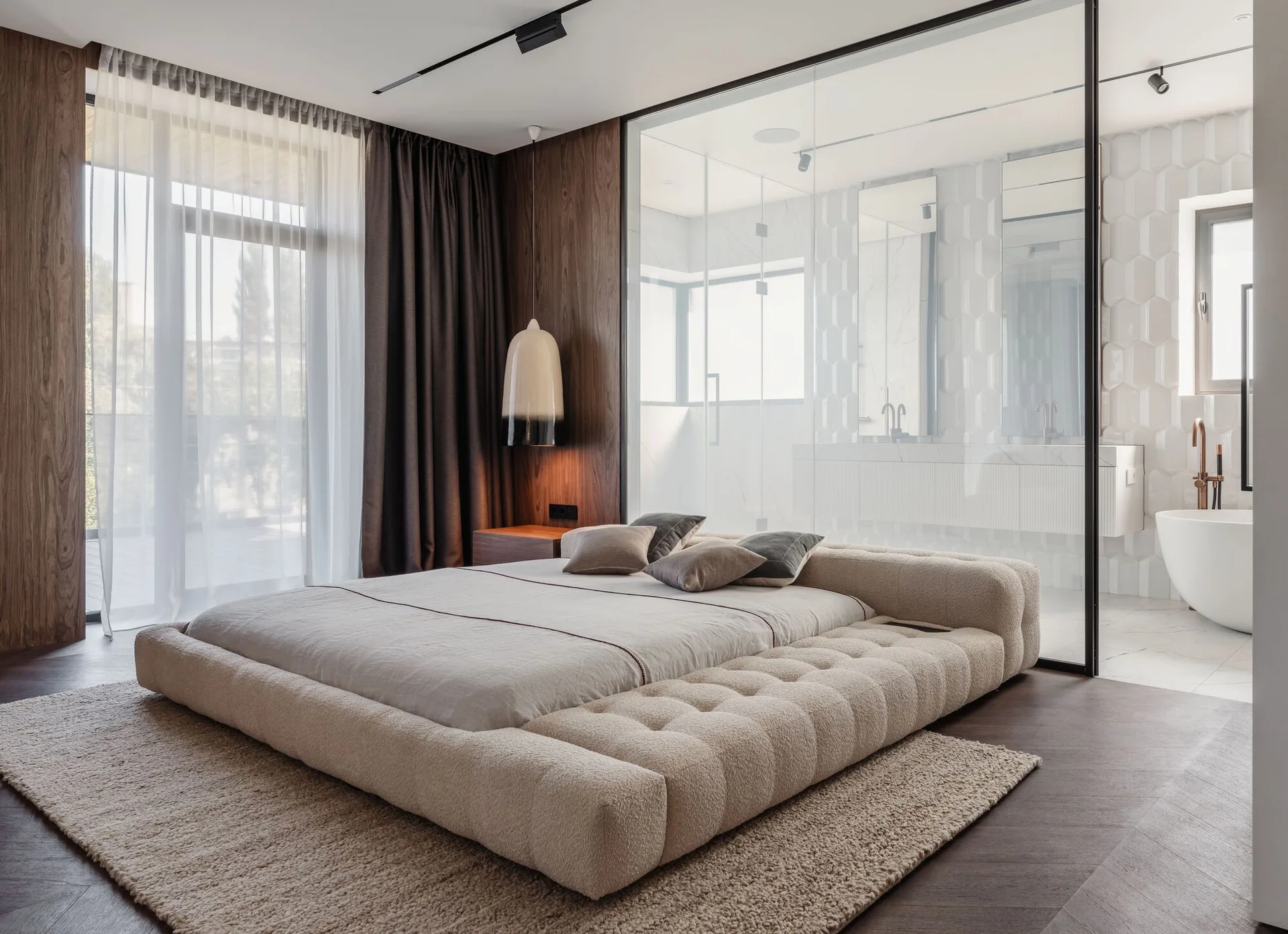 A low-profile tufted platform bed sits on a shag rug in a bedroom featuring dark wood paneling and a glass wall looking into a modern bathroom.