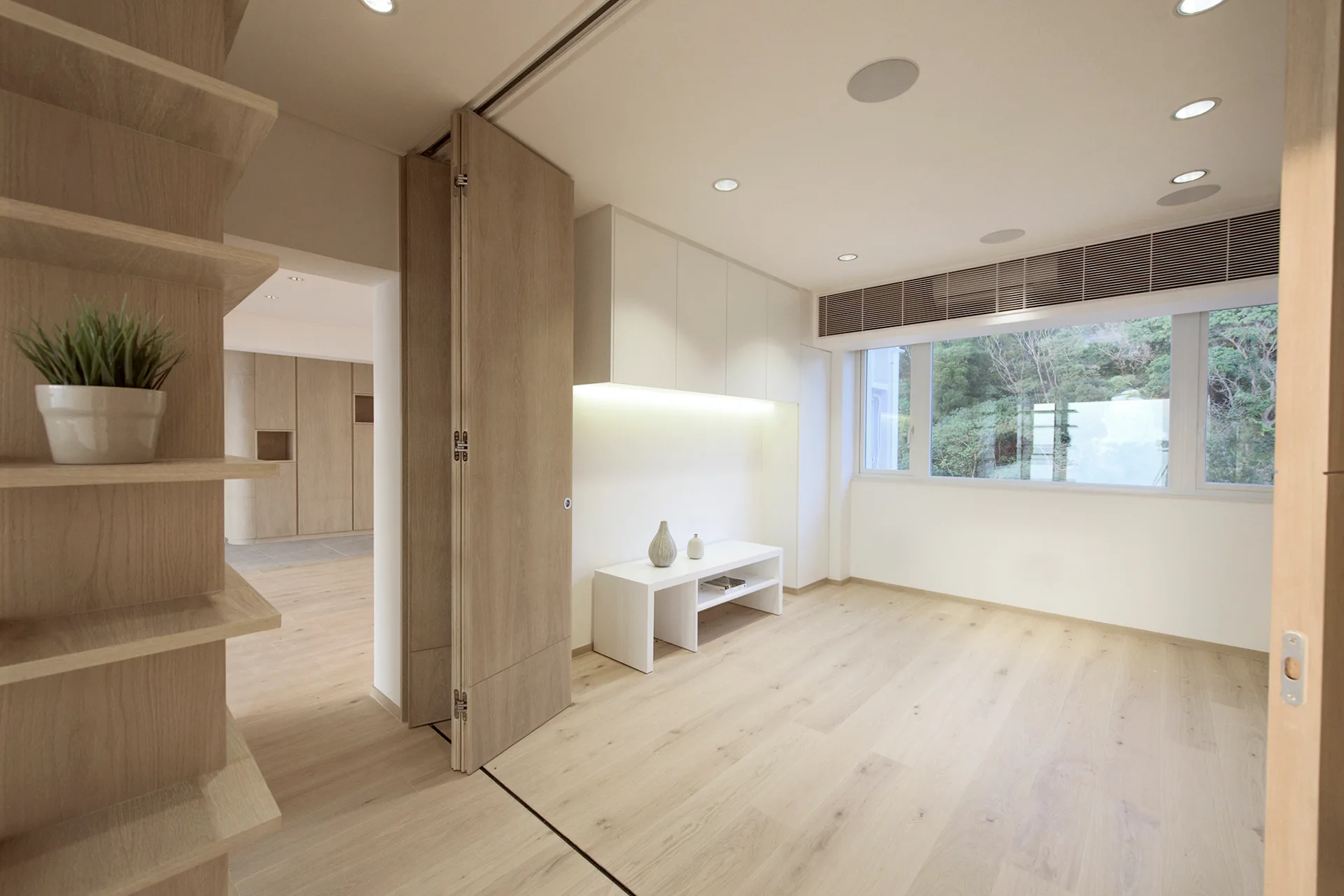 A minimalist living room features light wood floors, a tall bi-fold wooden door, built-in white cabinets, and a large window overlooking green trees.