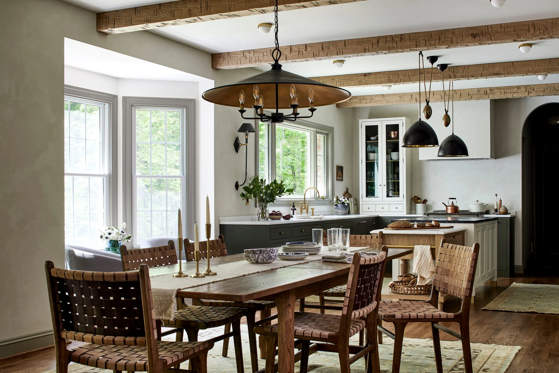 A wooden dining table with woven leather chairs sits under exposed ceiling beams, with a kitchen featuring dark cabinets in the background.
