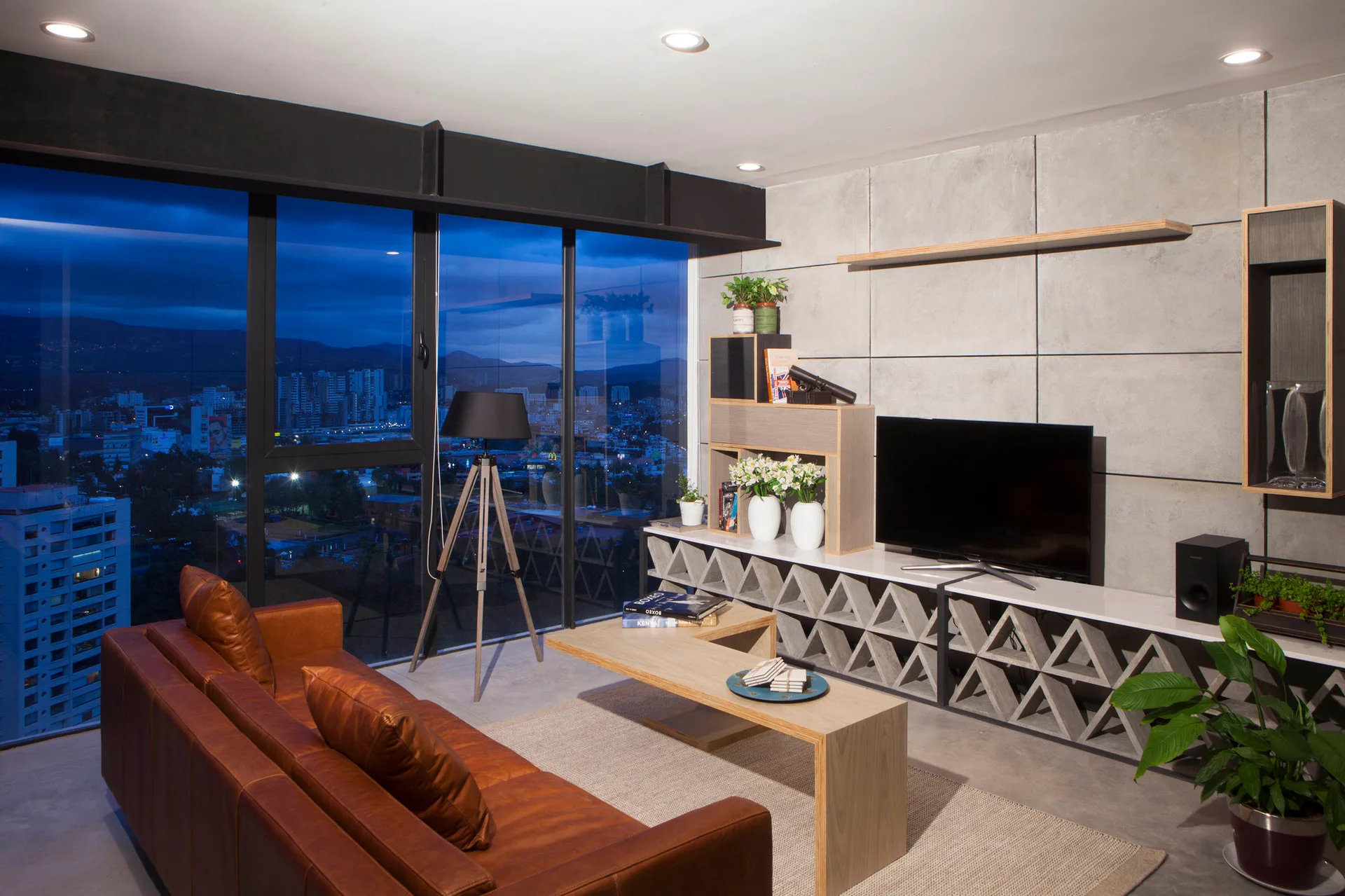 A modern living room features a brown leather sofa, a geometric concrete media console, and a floor-to-ceiling window showcasing a city night view.