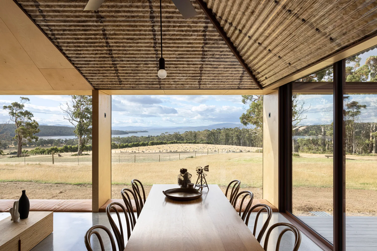 A minimalist dining room with a wooden table, bentwood chairs, a concrete floor, and a panoramic window overlooking a rural pasture.