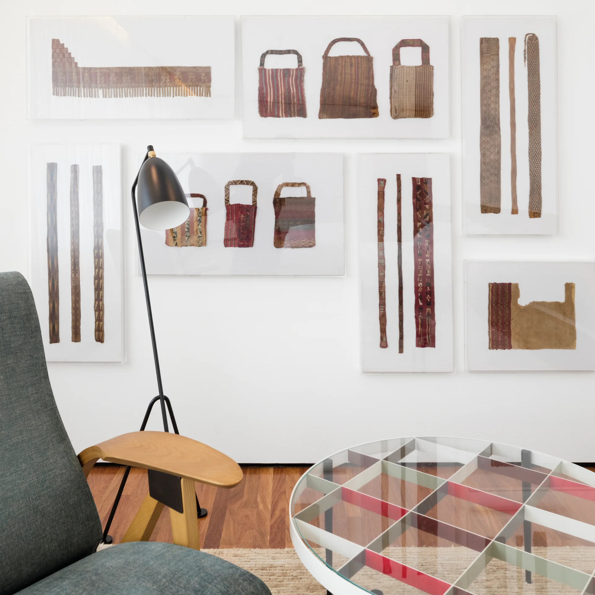 A contemporary living room features a gallery wall of framed textiles, a teal armchair, and a glass-top coffee table with a white grid frame.