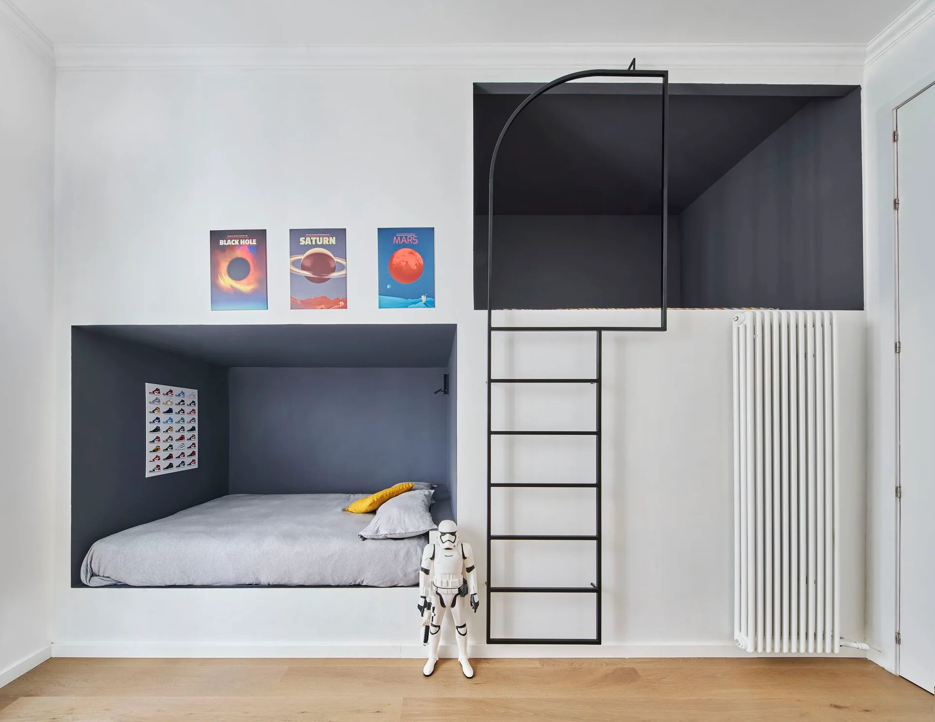 A kids bedroom features a recessed bed alcove painted dark grey, a black metal loft ladder, and space-themed posters on white walls.