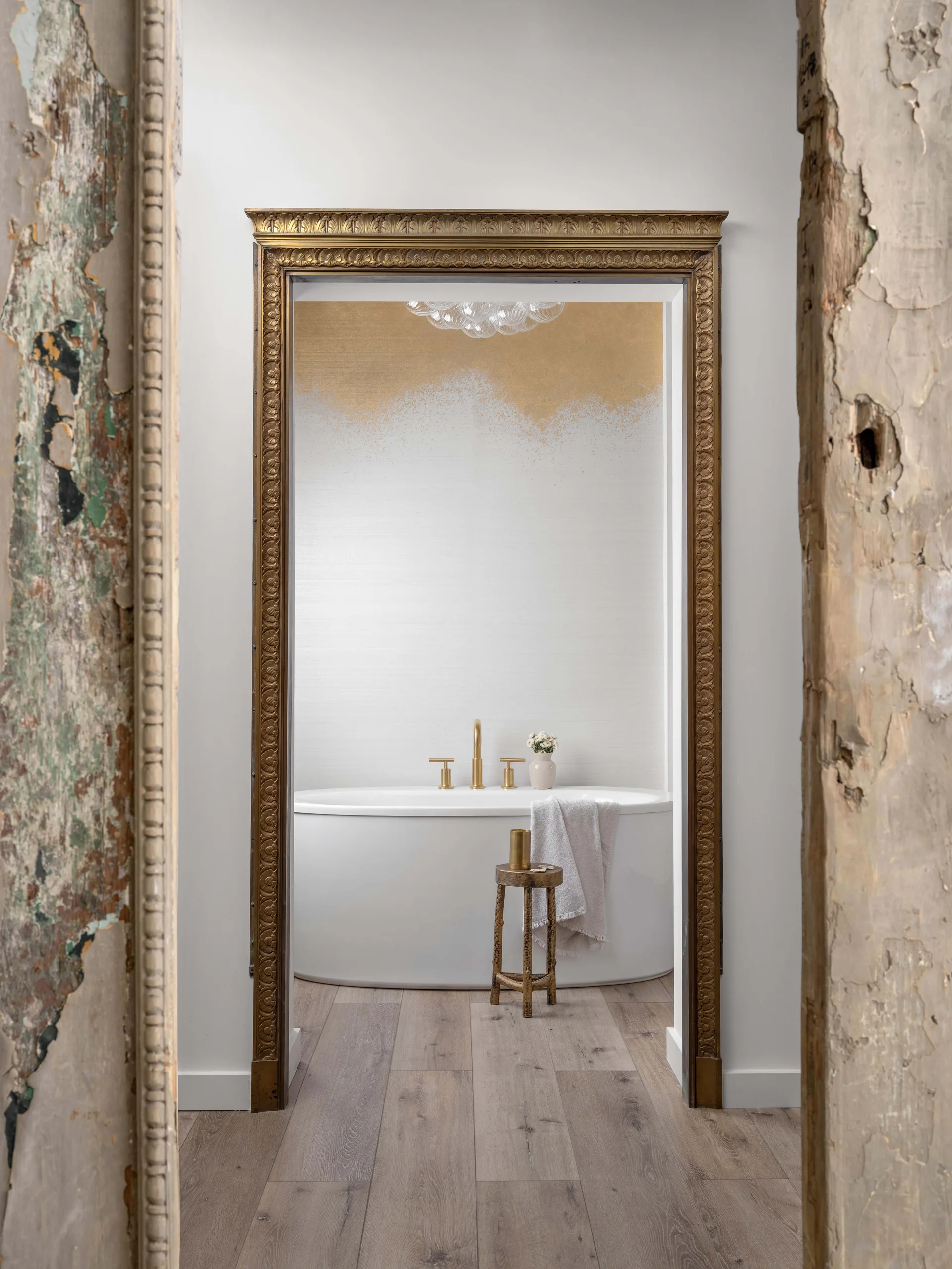 A white freestanding bathtub with gold fixtures is framed by an ornate gold archway, viewed through a distressed wooden doorway.