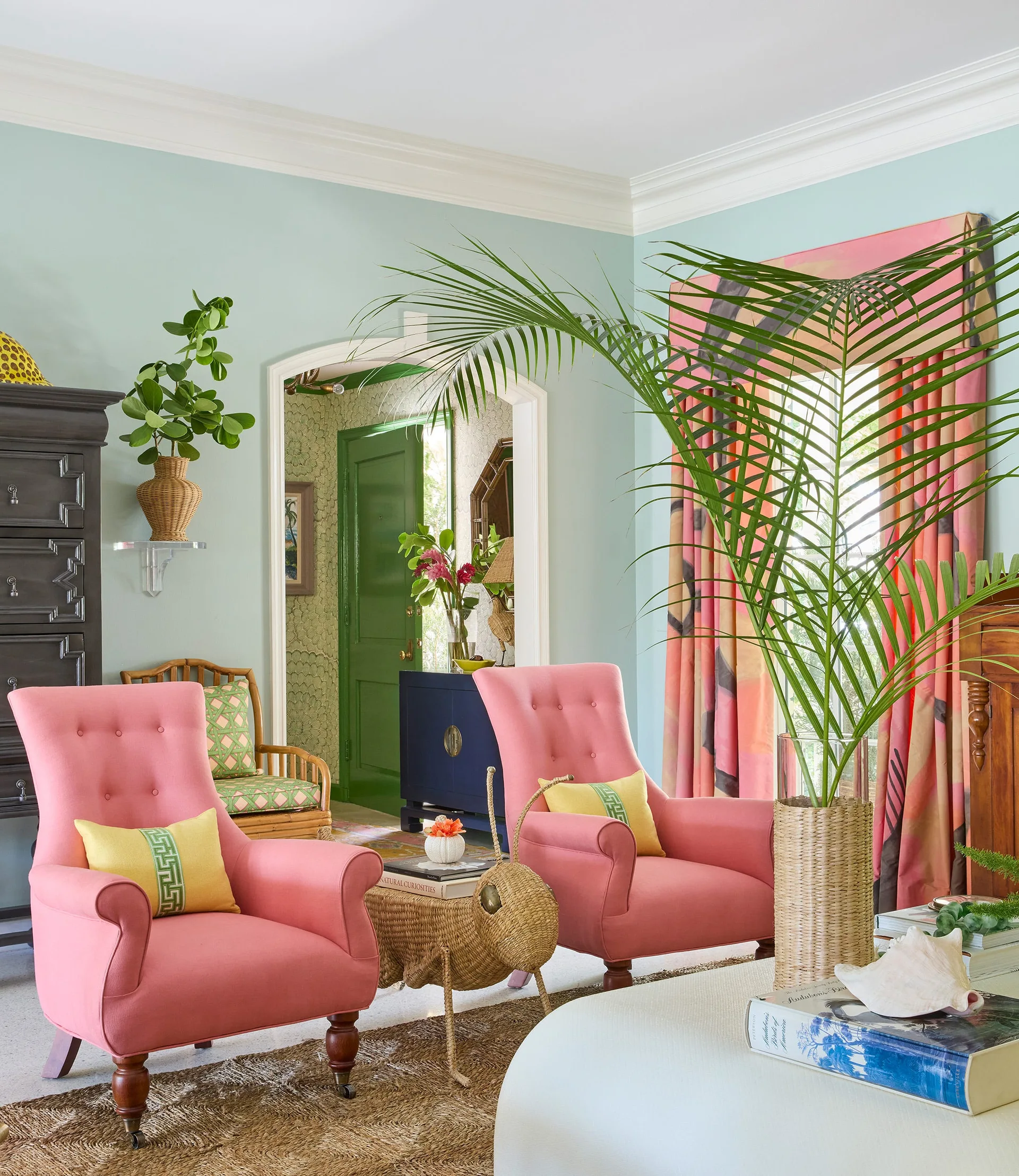 Two pink armchairs face a white coffee table in a light green living room featuring patterned pink curtains and a woven animal-shaped table.