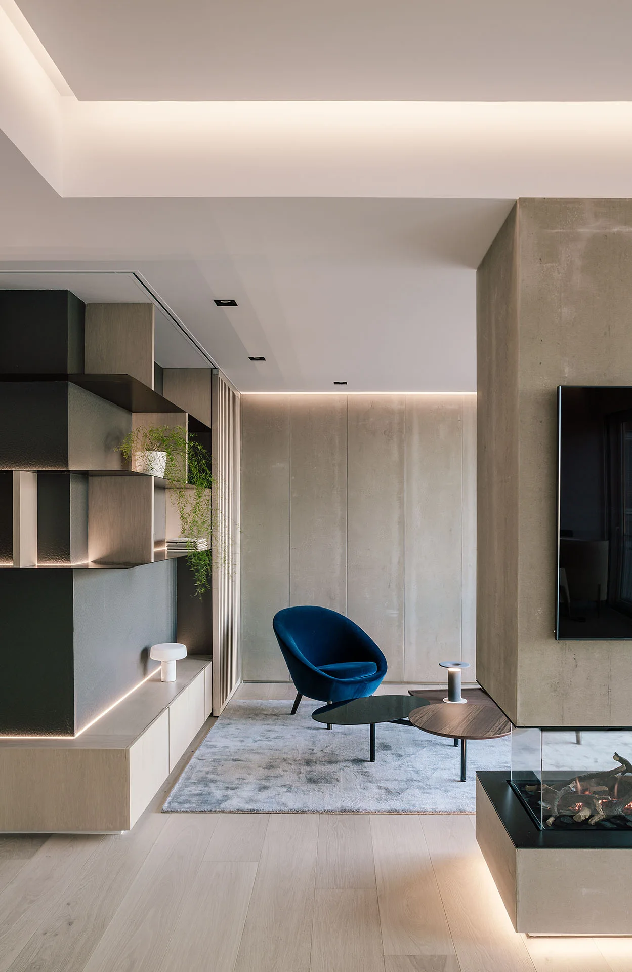 A modern living room features a blue velvet armchair, built-in wooden shelving with integrated lighting, and a concrete-finished fireplace.