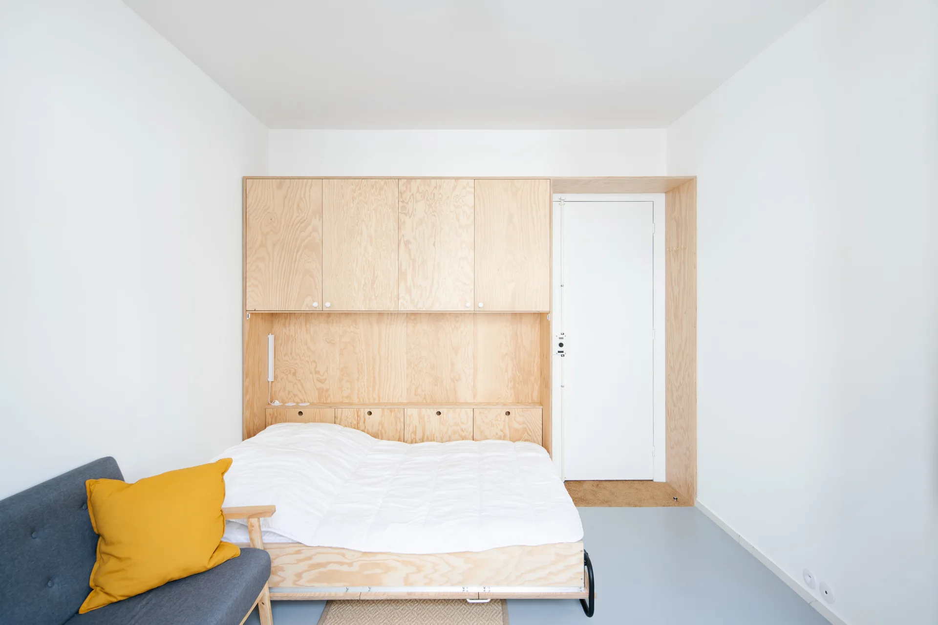 A minimalist bedroom featuring a bed integrated into a custom plywood storage unit with overhead cabinets next to a white door and grey sofa.