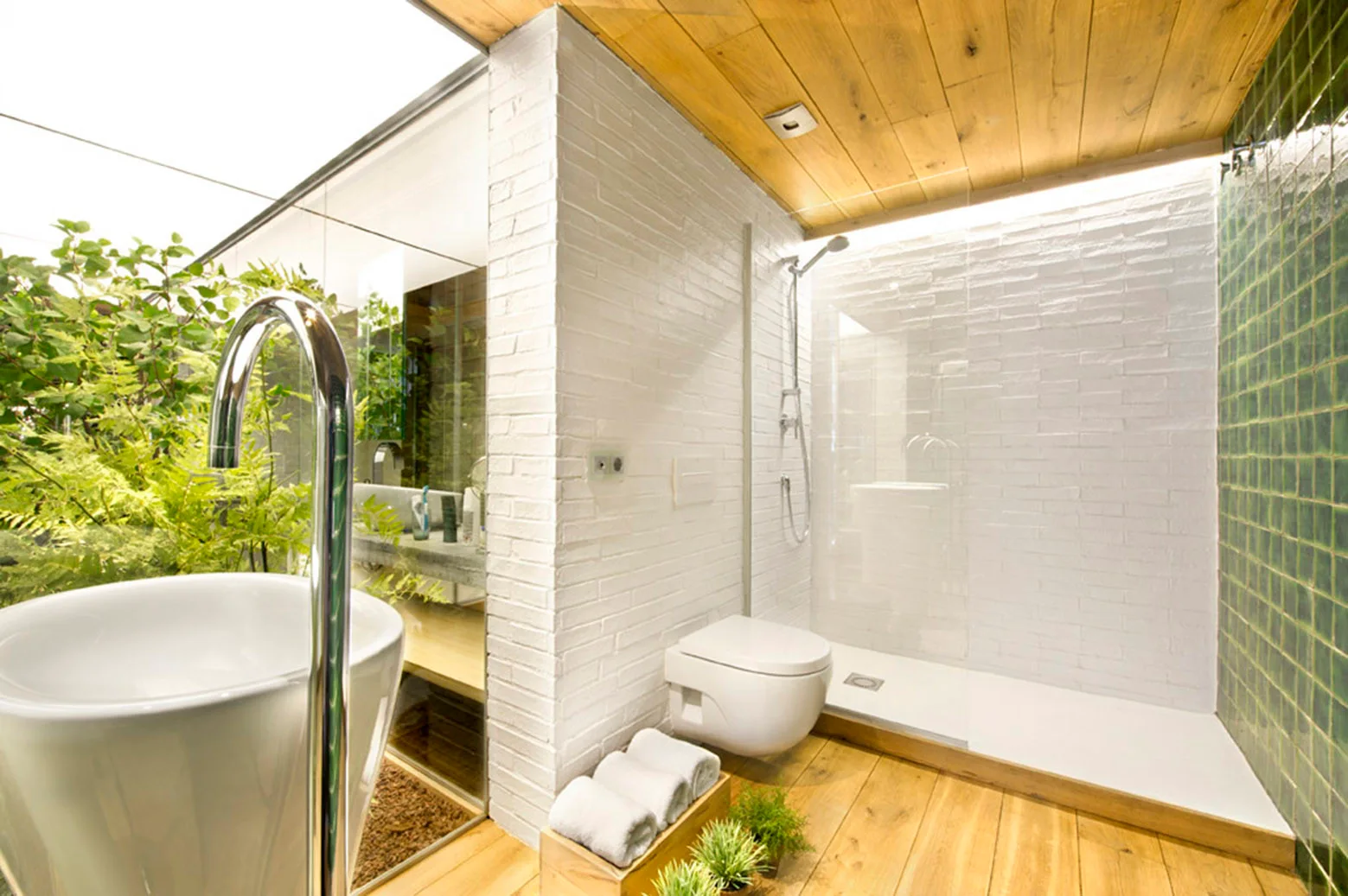 A modern bathroom features white brick walls, a wood ceiling and floor, a walk-in shower with green tiles, and a view of a lush garden.