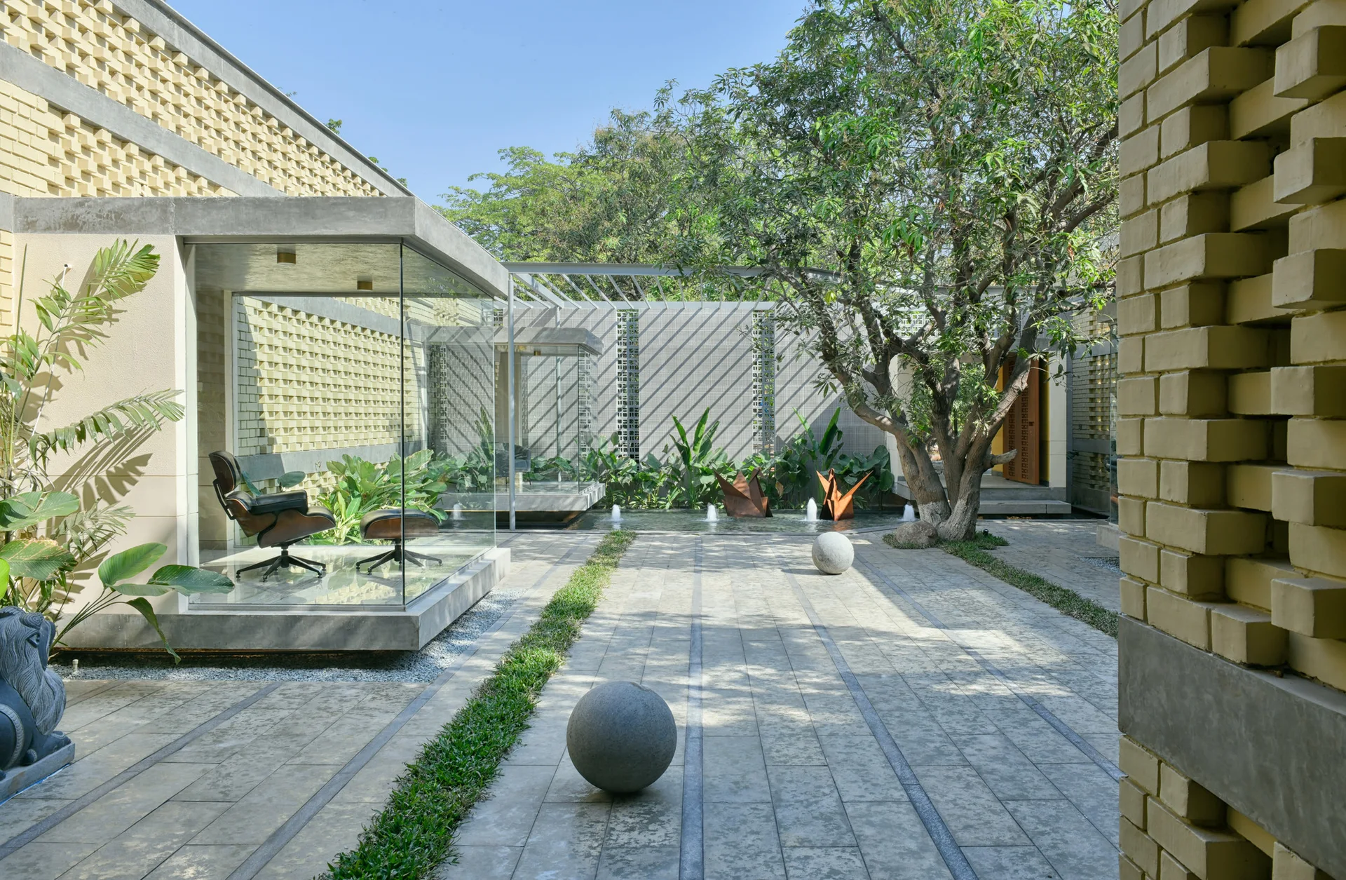A modern outdoor courtyard featuring a glass-enclosed lounge chair pavilion, textured yellow brick walls, stone pavers, and spherical sculptures.