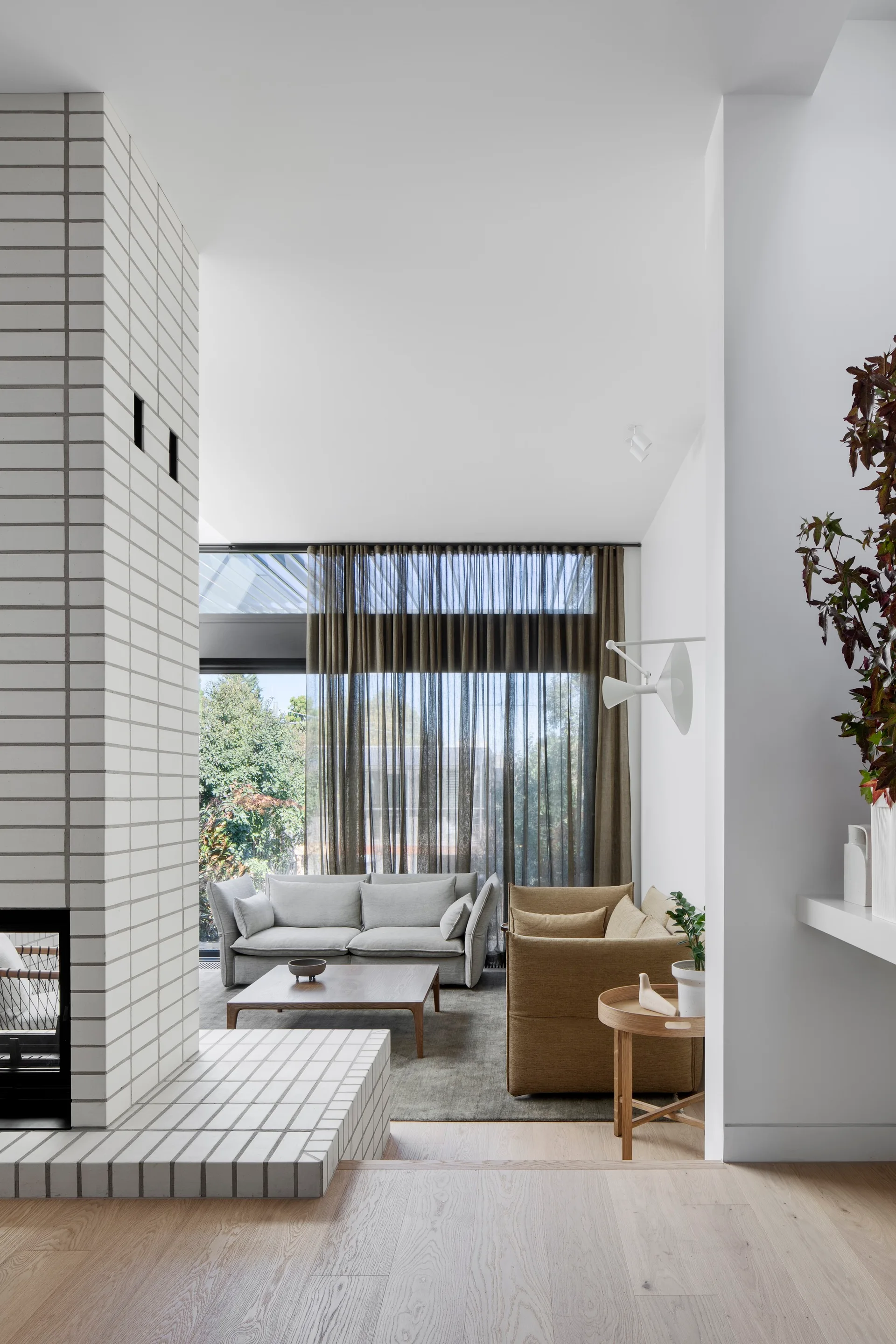 A minimalist living room featuring a tall white brick fireplace, gray and tan sofas, a wood coffee table, and sheer floor-to-ceiling curtains.