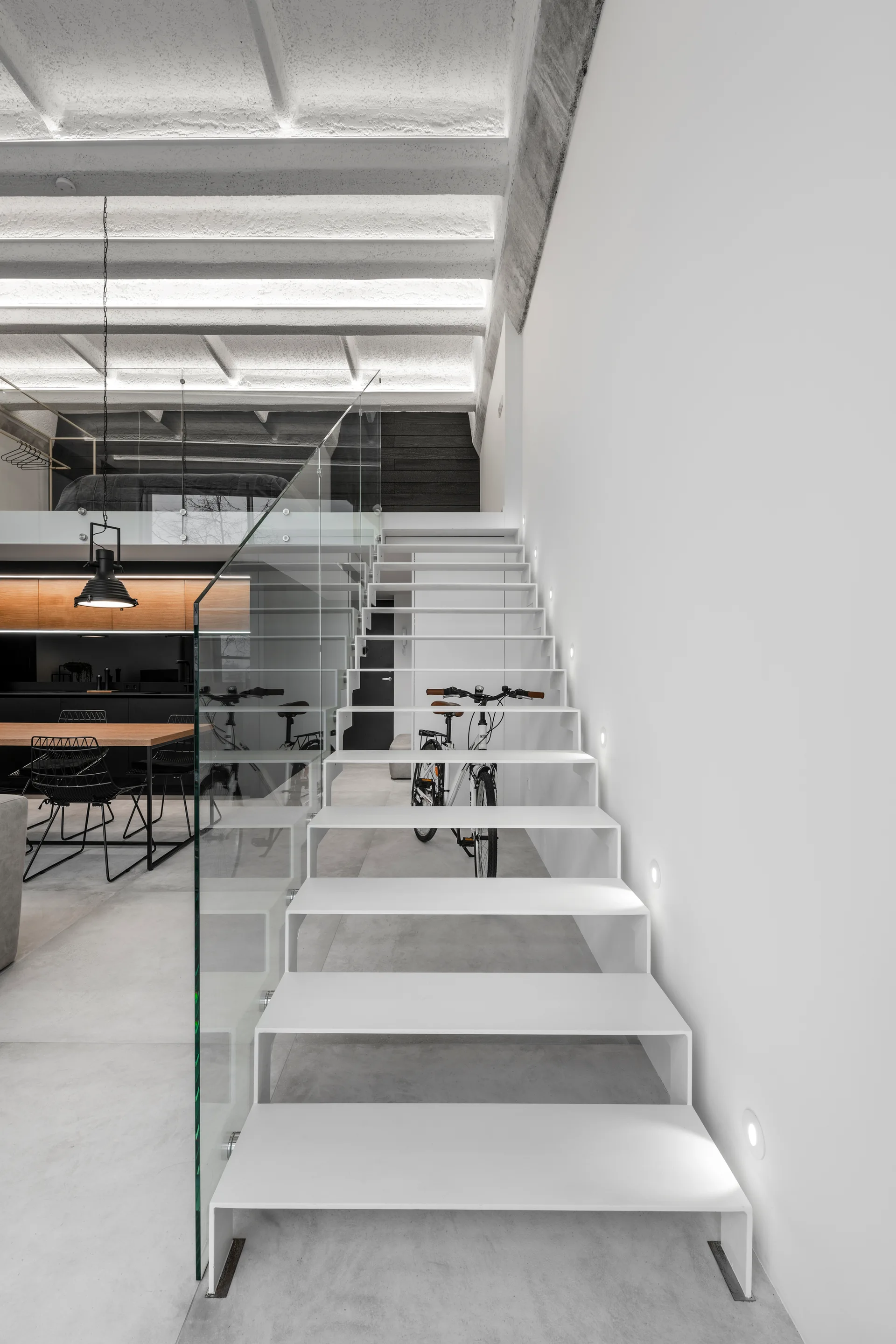 A modern white folded metal staircase features a glass railing and recessed wall lights, set against a concrete floor and exposed beamed ceiling.