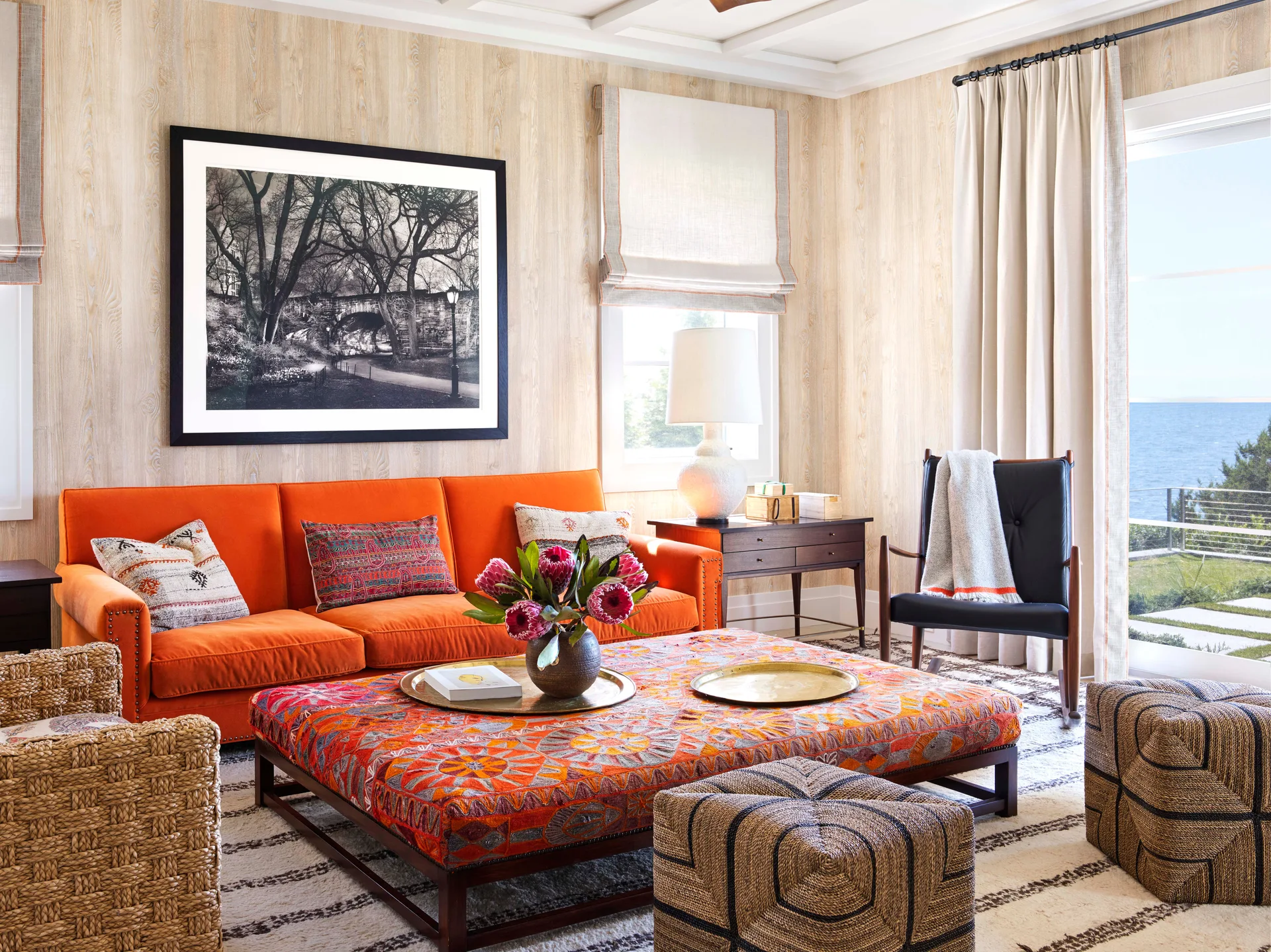 A living room with an orange sofa, a large patterned fabric ottoman, woven stools, wood-patterned wallpaper, and a window showing an ocean view.