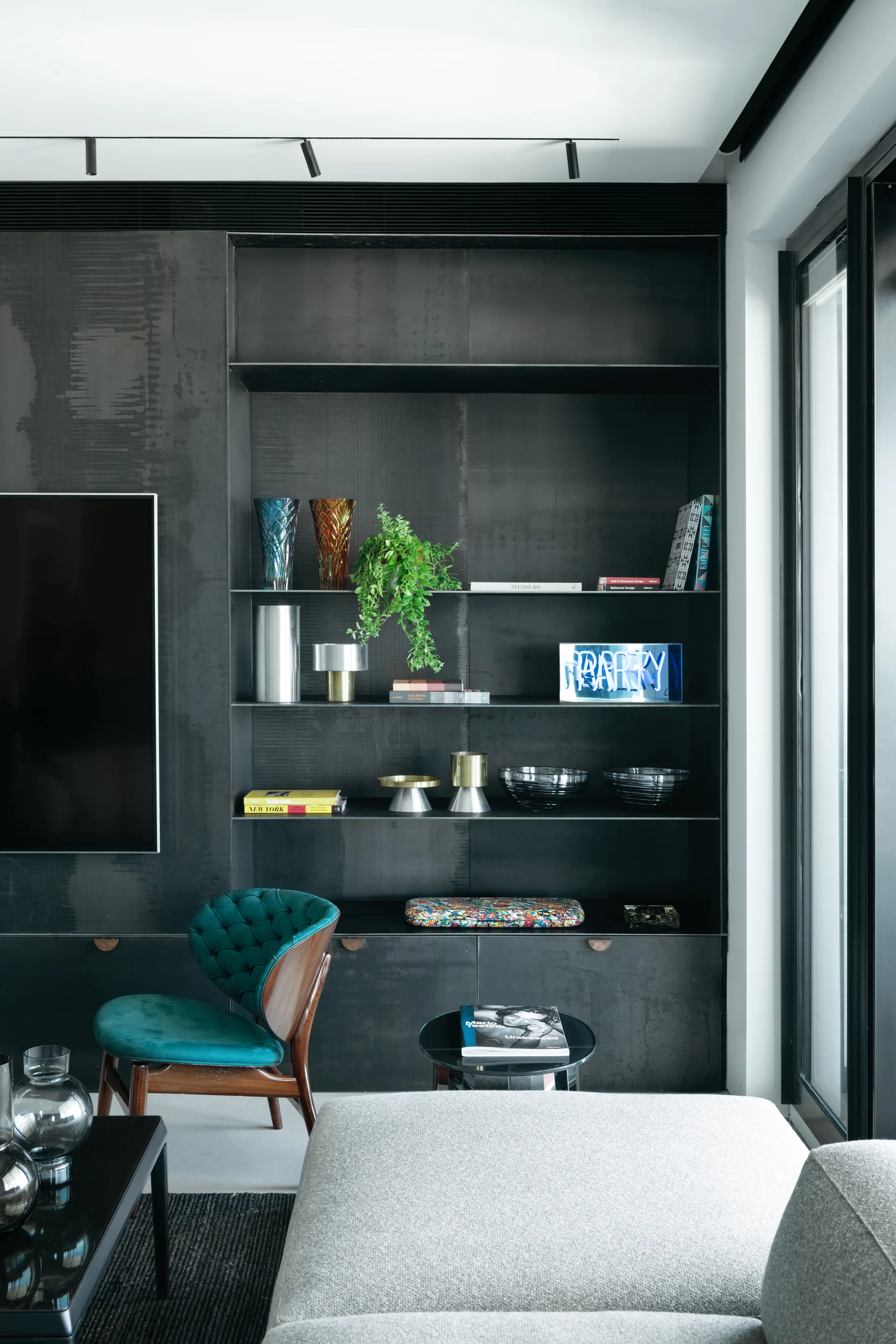 A dark charcoal metal built-in shelving unit displays books and colorful glass vases behind a teal velvet armchair and grey sofa.