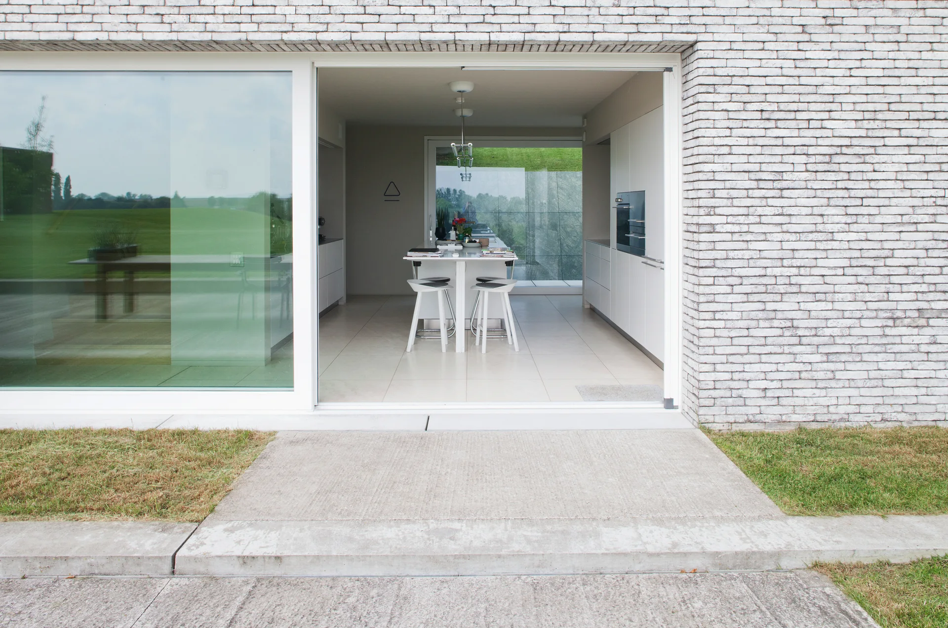 A modern minimalist kitchen with a central island and white stools is seen through an open sliding glass door of a white brick building.