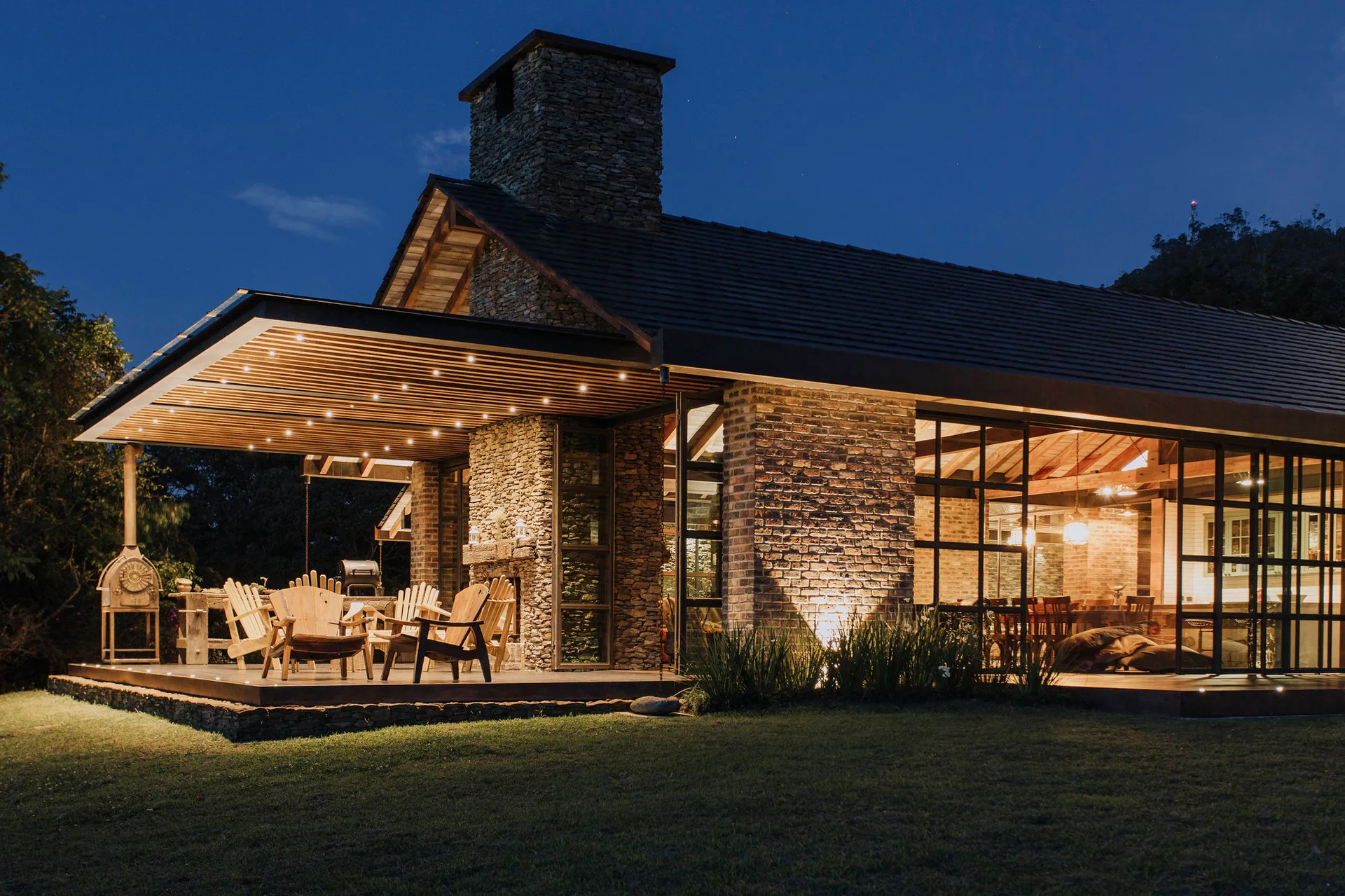 A rustic covered patio at night with wooden Adirondack chairs, a stone fireplace, and light shining through large metal-framed windows.