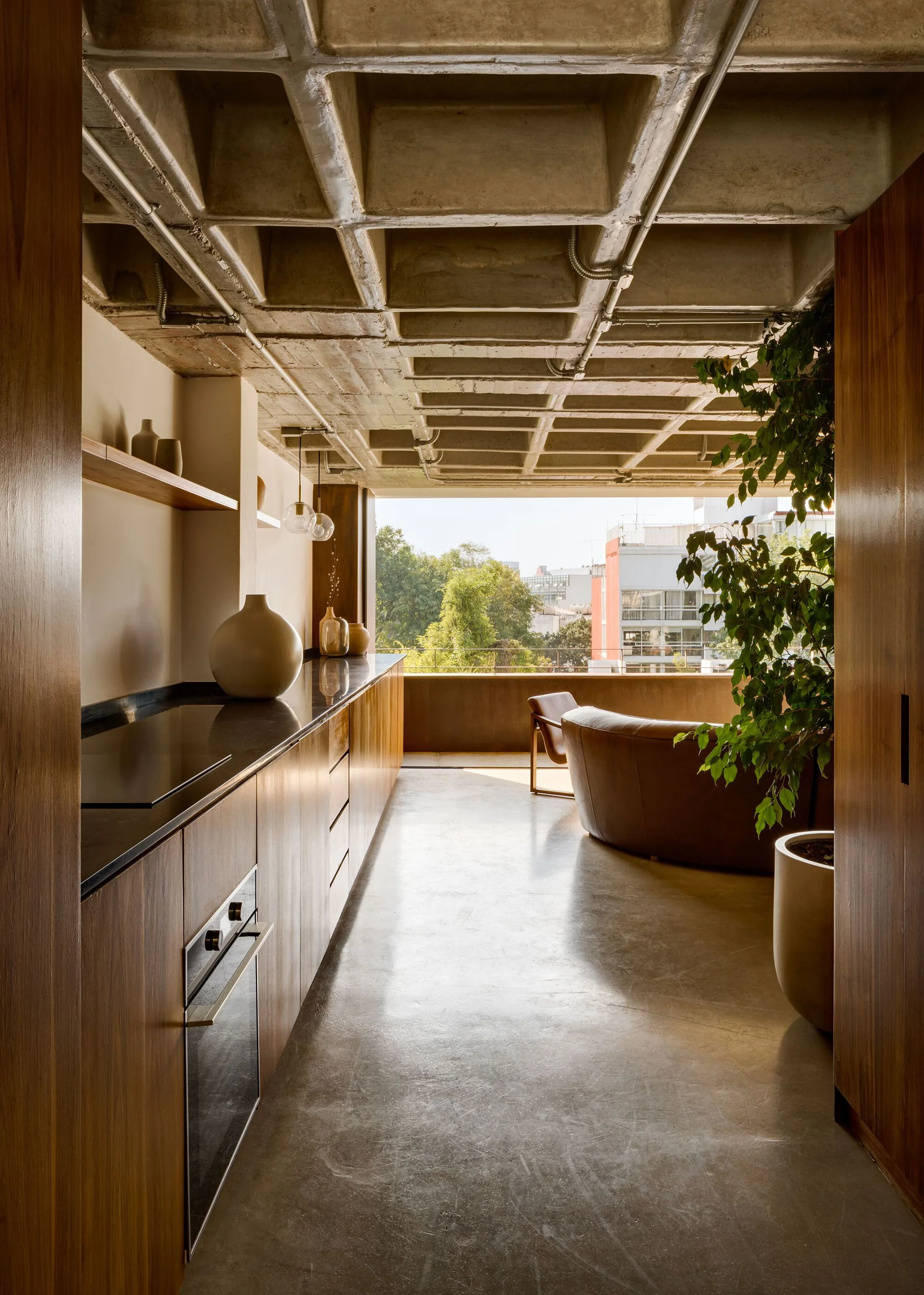 A modern kitchen features long wood cabinets, a polished concrete floor, and an exposed concrete waffle ceiling leading to an outdoor balcony.
