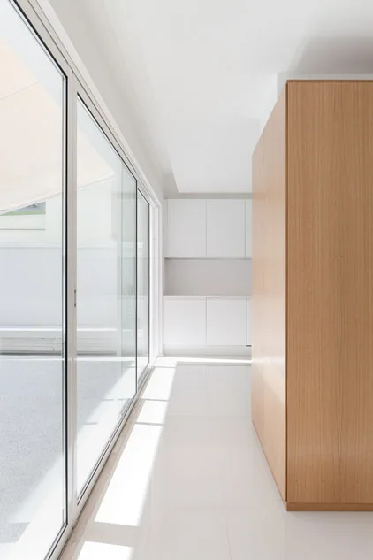 High-contrast minimalist interior featuring a tall light-wood cabinet panel, glossy white floors, and expansive sliding glass doors leading outside.