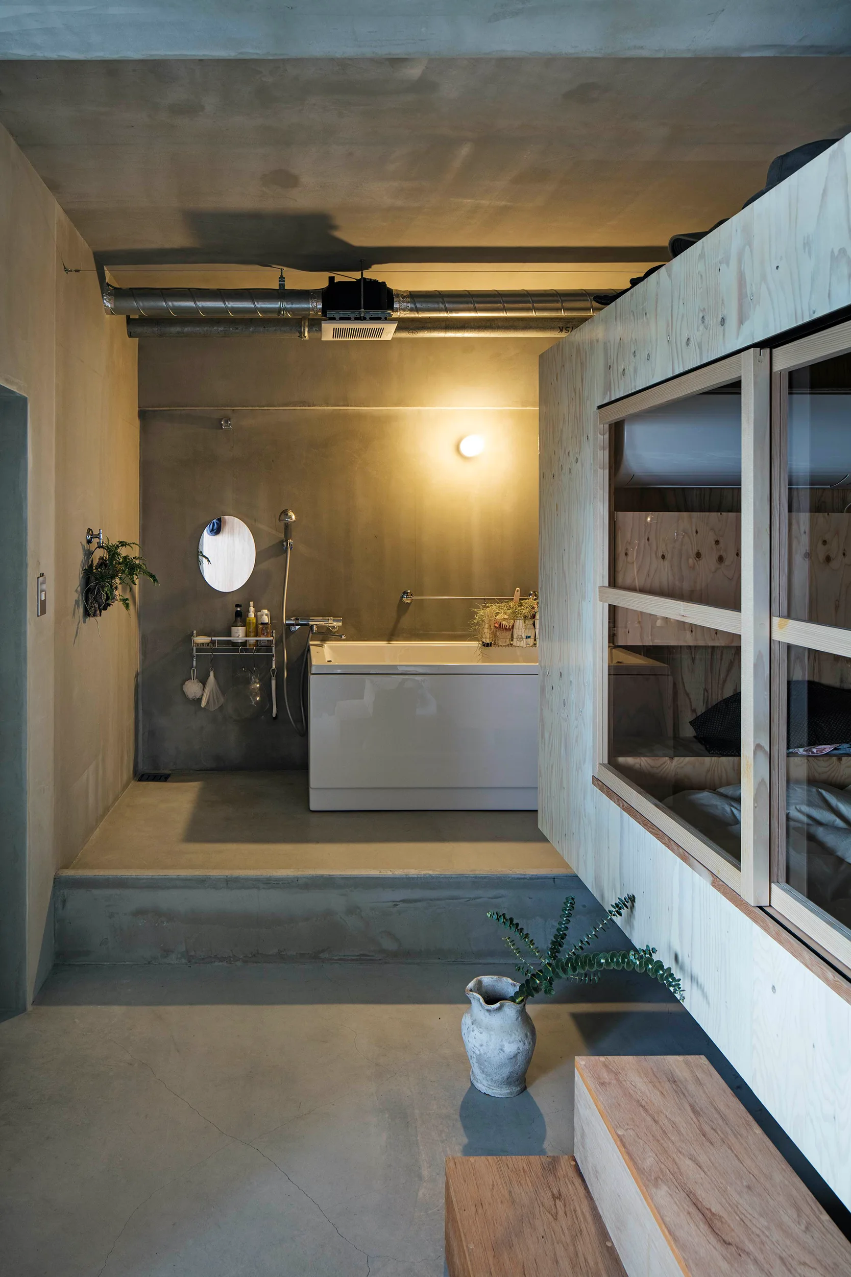 A modern industrial bathroom features a white bathtub on a raised concrete platform next to a raw plywood partition wall.