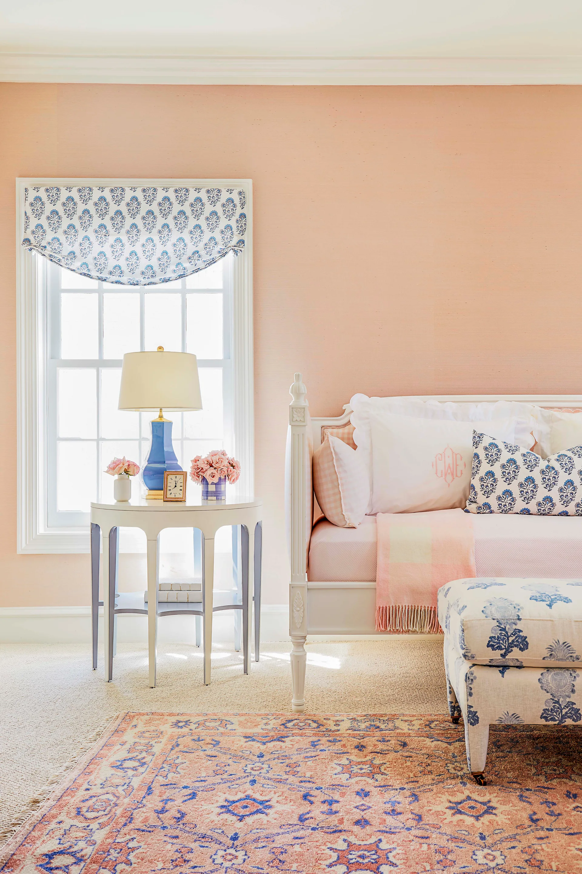 A white daybed with blue patterned pillows sits next to a round nightstand with a blue lamp under a window with a matching patterned valance.