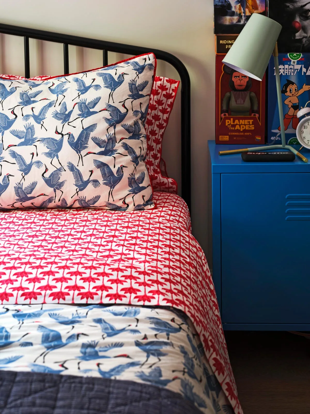 A black metal bed frame with crane-print pillows and red patterned sheets next to a bright blue metal locker-style nightstand.